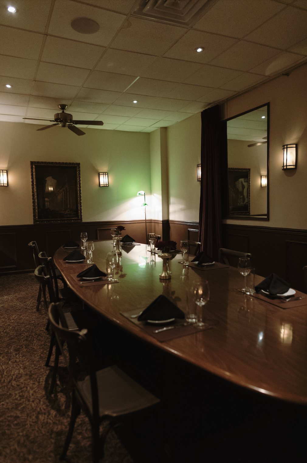 A dimly lit dining room with a long oval table set with black napkins, glasses, and small floral centerpieces. The room has wall sconces, a mirror, and a framed picture on the wall, with a ceiling fan and a floor lamp providing soft lighting.