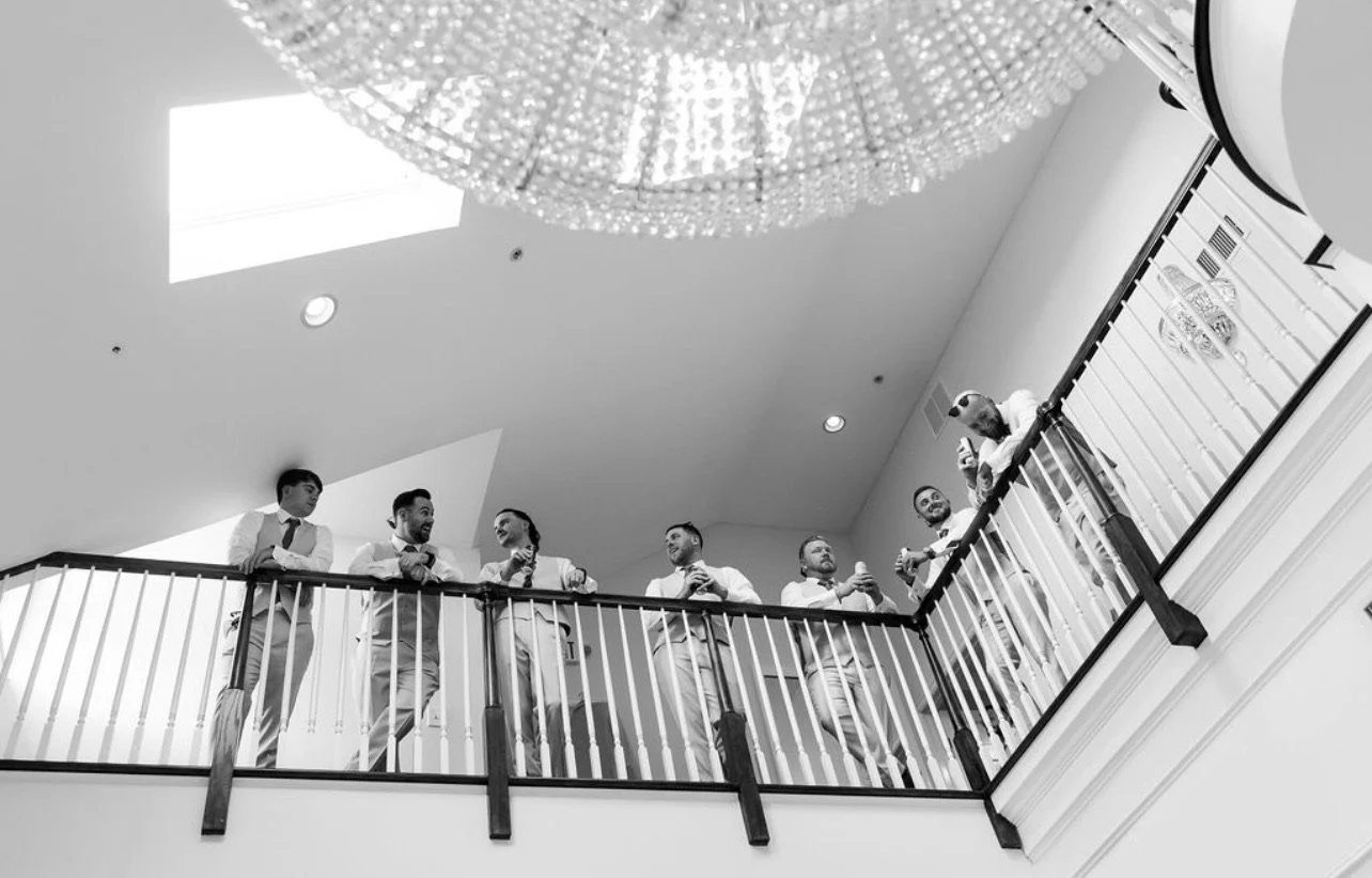 Group of men dressed in formal attire standing on an upper-level balcony, engaging in conversation and some holding drinks, overlooking a chandelier and a lower level.