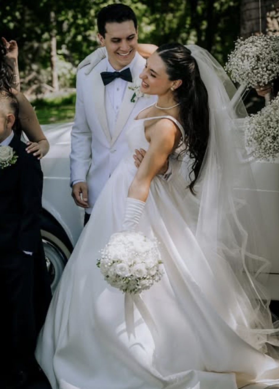 A bride and groom in wedding attire smiling and looking at each other outside, with a young boy in a suit nearby, and decorated with flowers and greenery.
