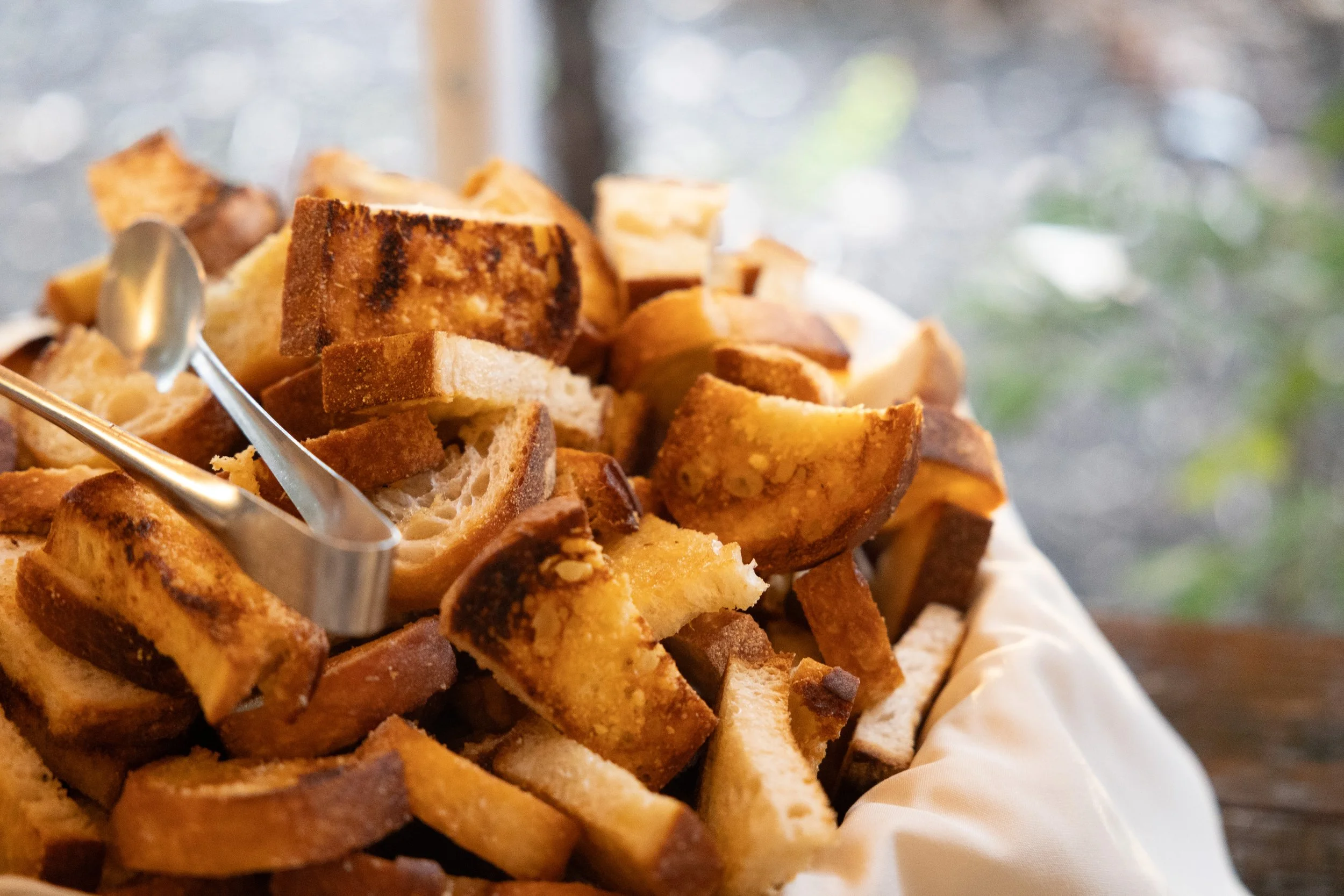 A basket of toasted bread slices on a white cloth, with tongs resting on top. The background is blurred with hints of greenery.