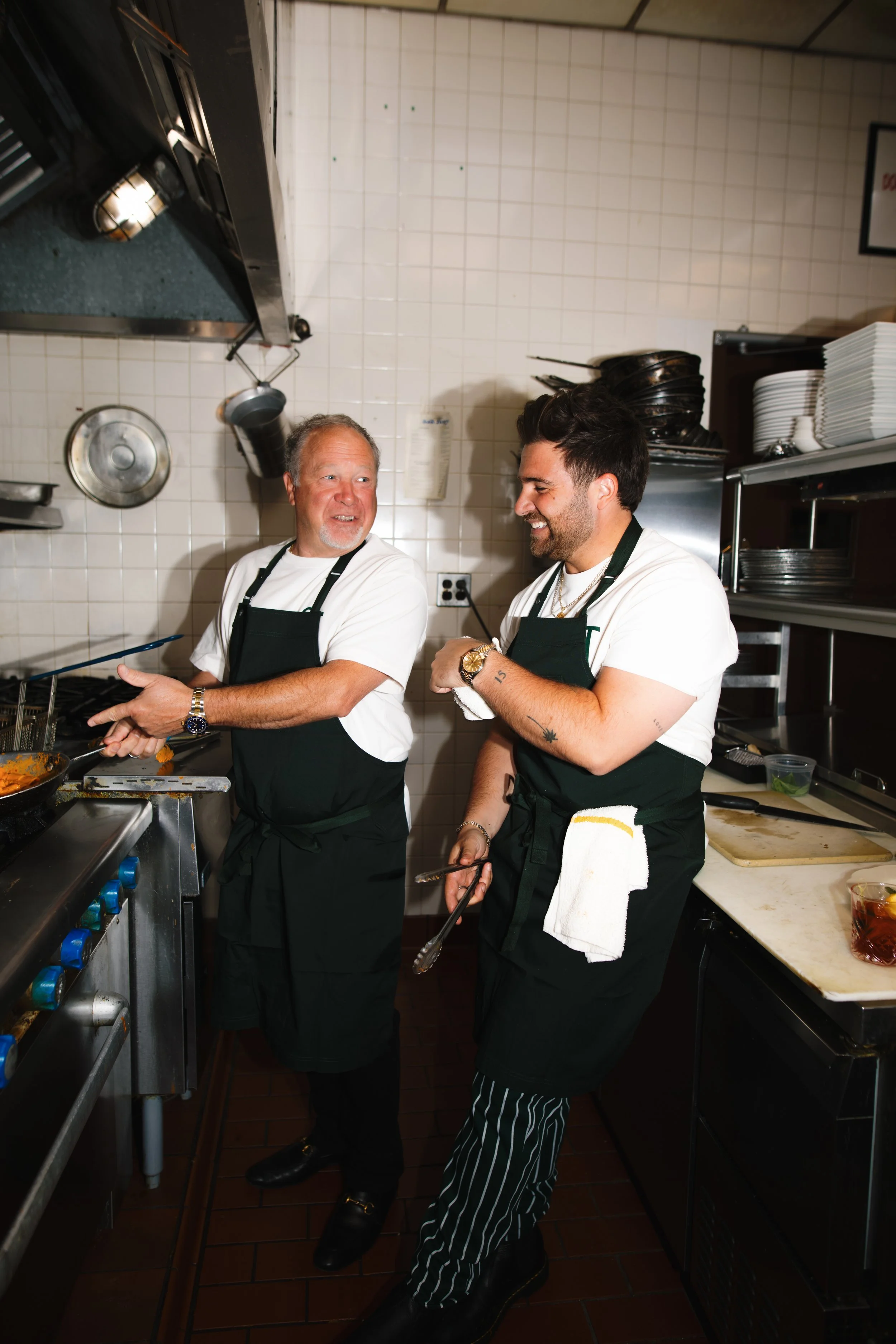 Two male chefs in a restaurant kitchen smiling and talking; one is cooking while the other is standing beside him with tongs in hand.