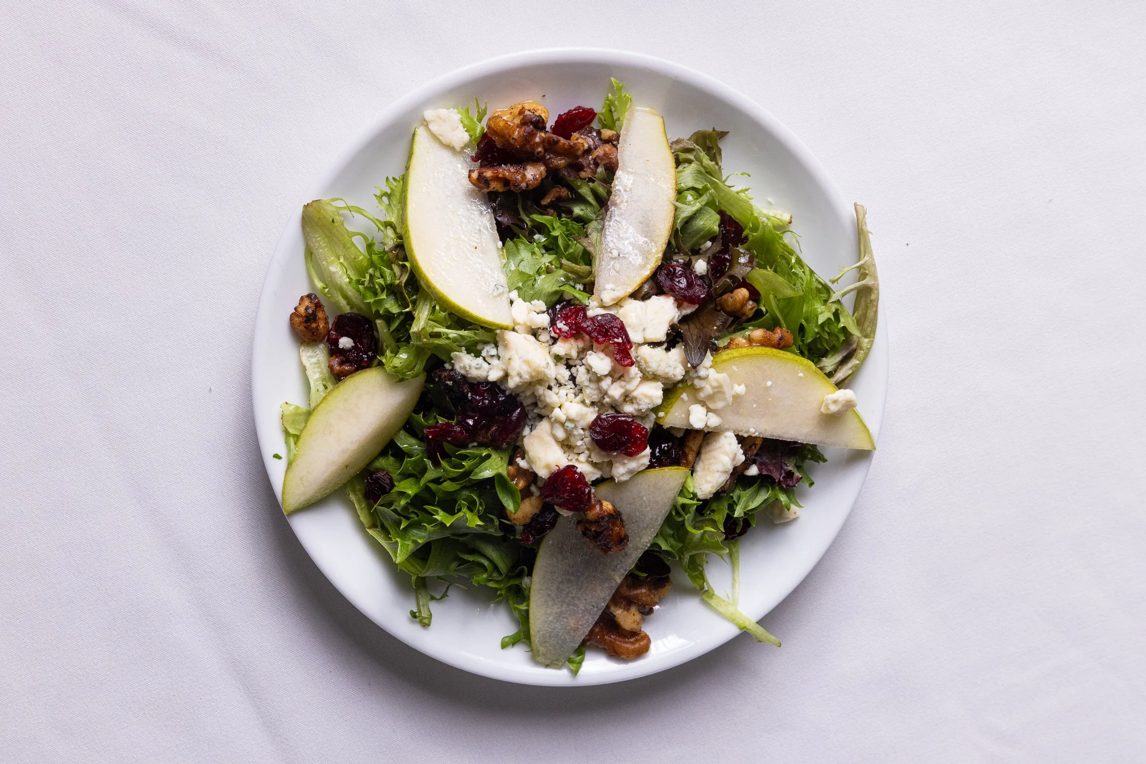 Fresh mixed green salad with pear slices, crumbled feta cheese, dried cranberries, and walnuts on a white plate.