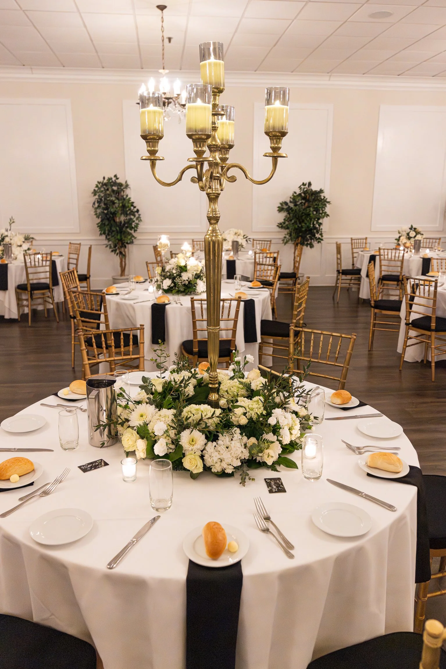 Elegant banquet table with a gold candelabrum centerpiece, surrounded by white floral arrangements, set for a formal event with plates, silverware, bread rolls, and candles, in a decorated banquet hall.