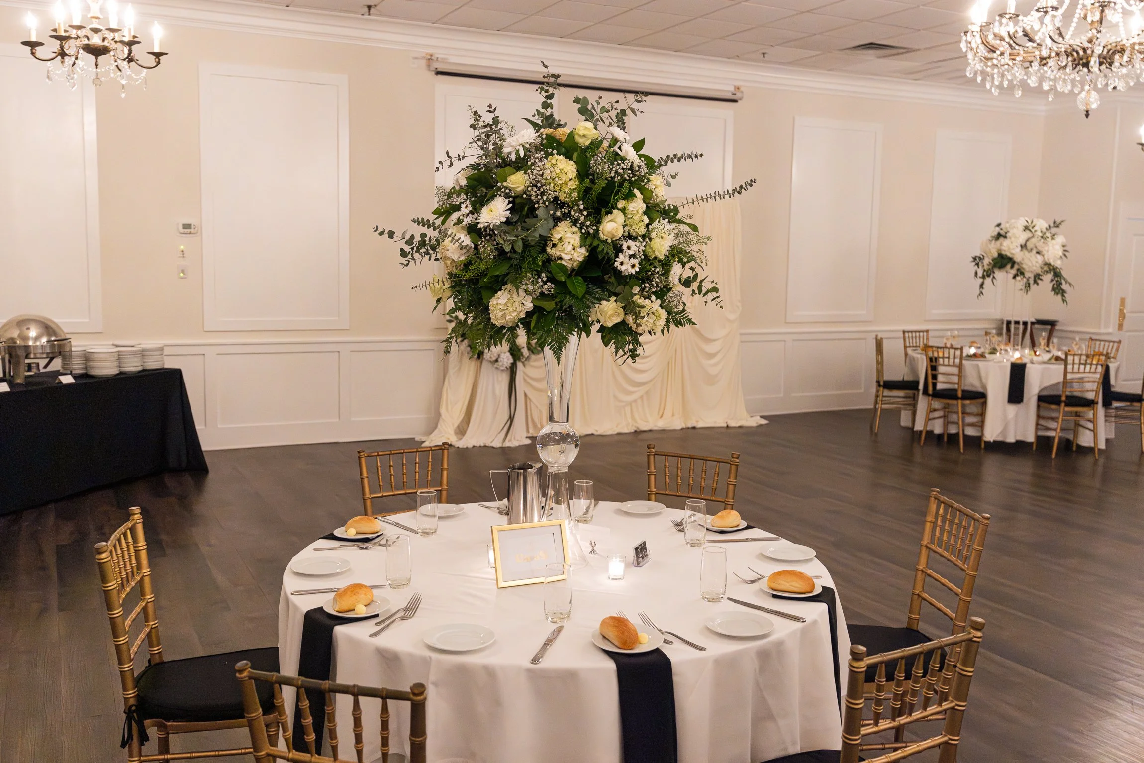 Elegant banquet hall with round table set for a formal event, decorated with a tall floral centerpiece of white flowers and greenery, surrounded by gold chairs, with a black tablecloth buffet station and additional tables with similar decorations in the background