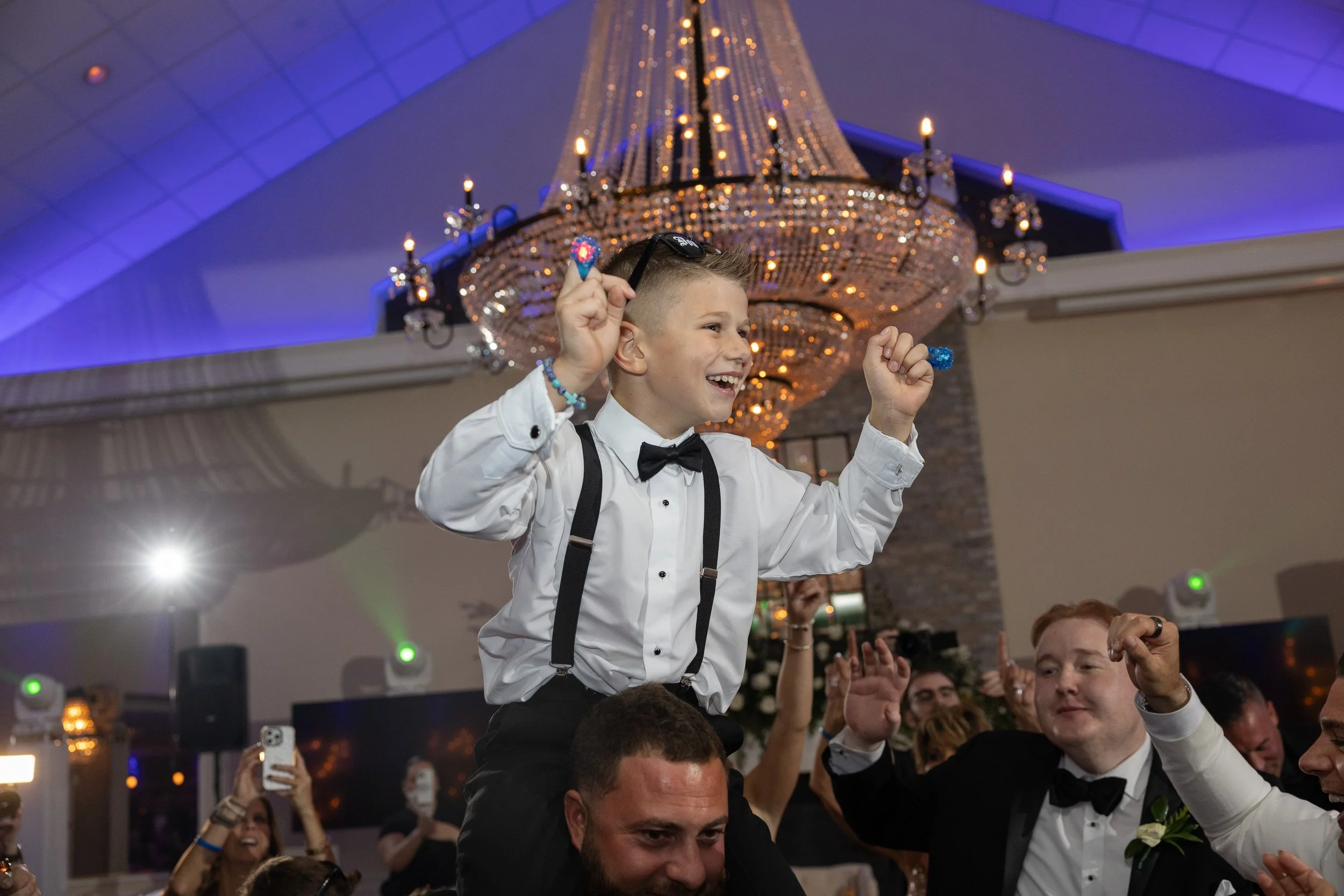 A young boy in a tuxedo and bow tie is riding on someone's shoulders at a celebration, smiling and raising his fists in excitement. People around him are dressed in formal attire, with some taking photos. The setting is an elegant event with a large 