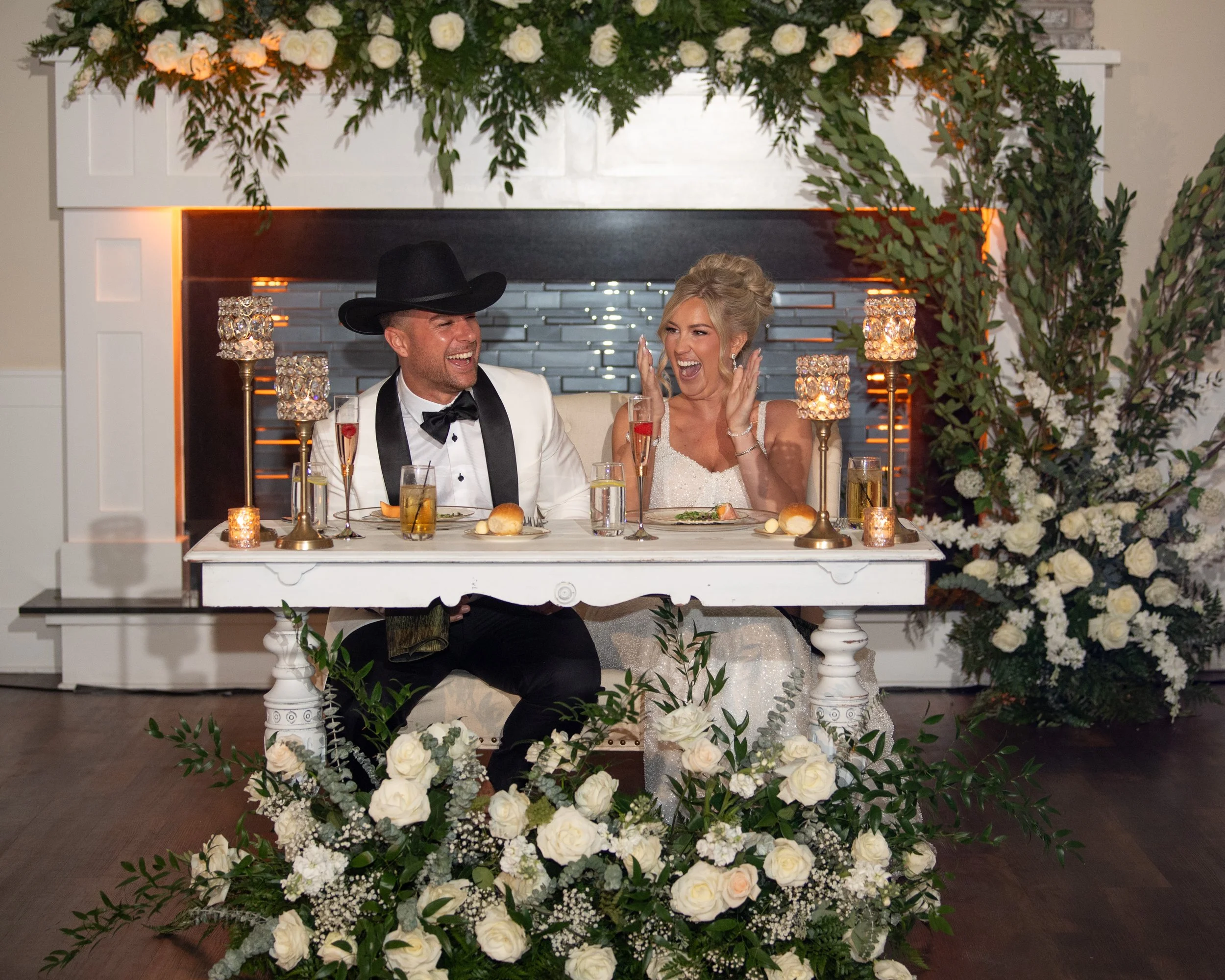 A bride and groom are seated at a table during their wedding reception, surrounded by white flowers and candles, sharing a joyful moment.