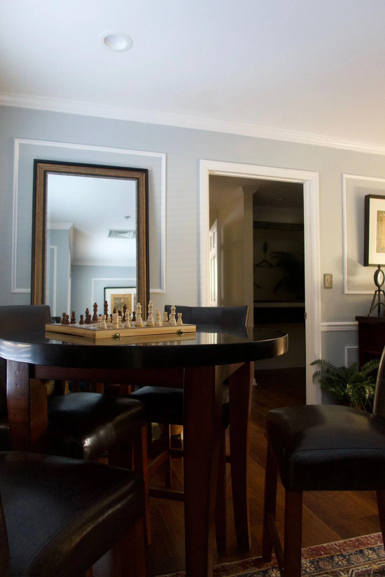 A dining room with a round black table, four black cushioned chairs, a chessboard with chess pieces on the table, a large mirror on the wall, and a doorway leading to a walk-in closet. The walls are painted light gray with white trim and crown moldin