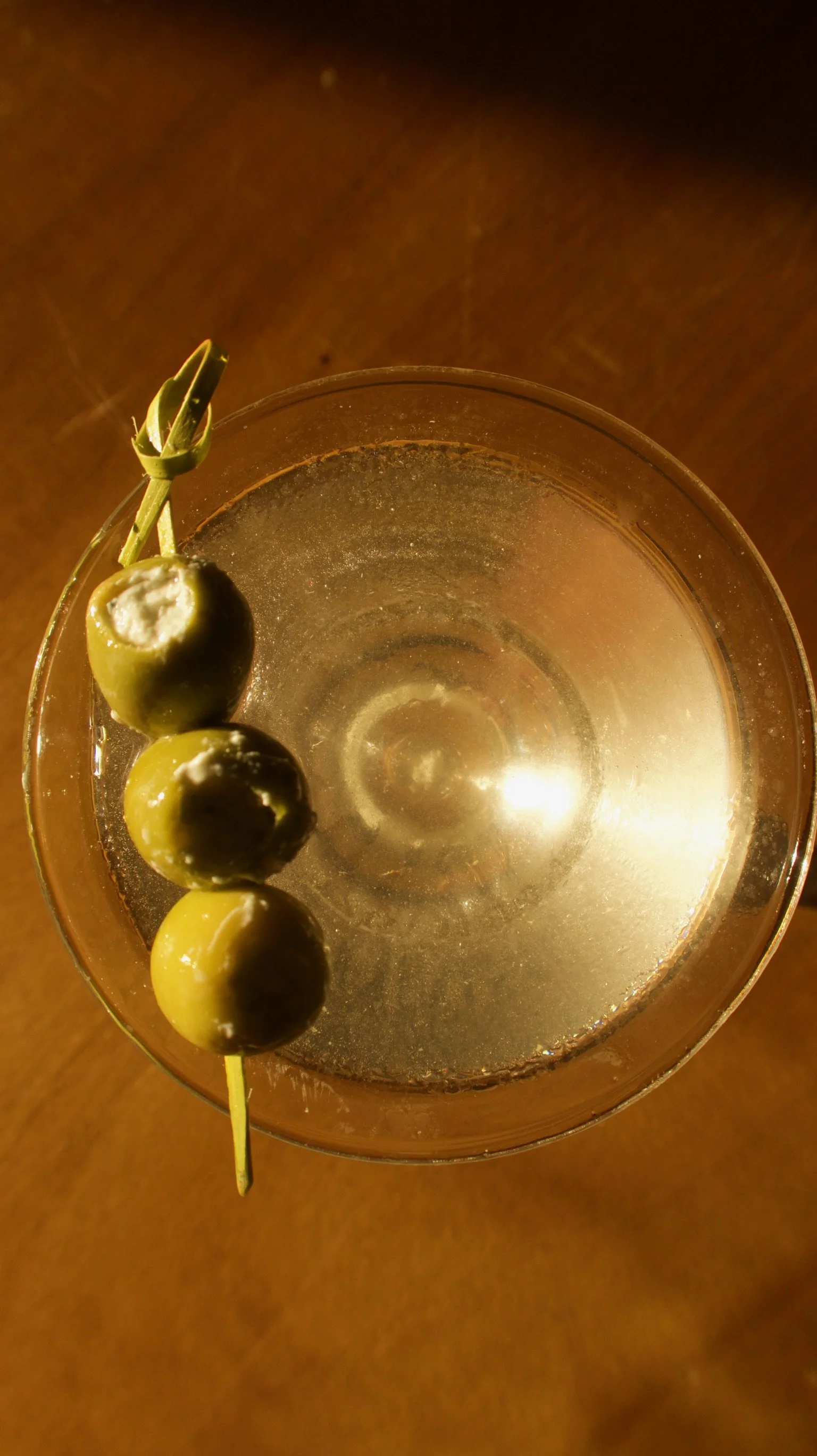 A glass of clear drink with a skewer of three green olives garnished with a small amount of white cheese or filling, placed on a wooden surface.