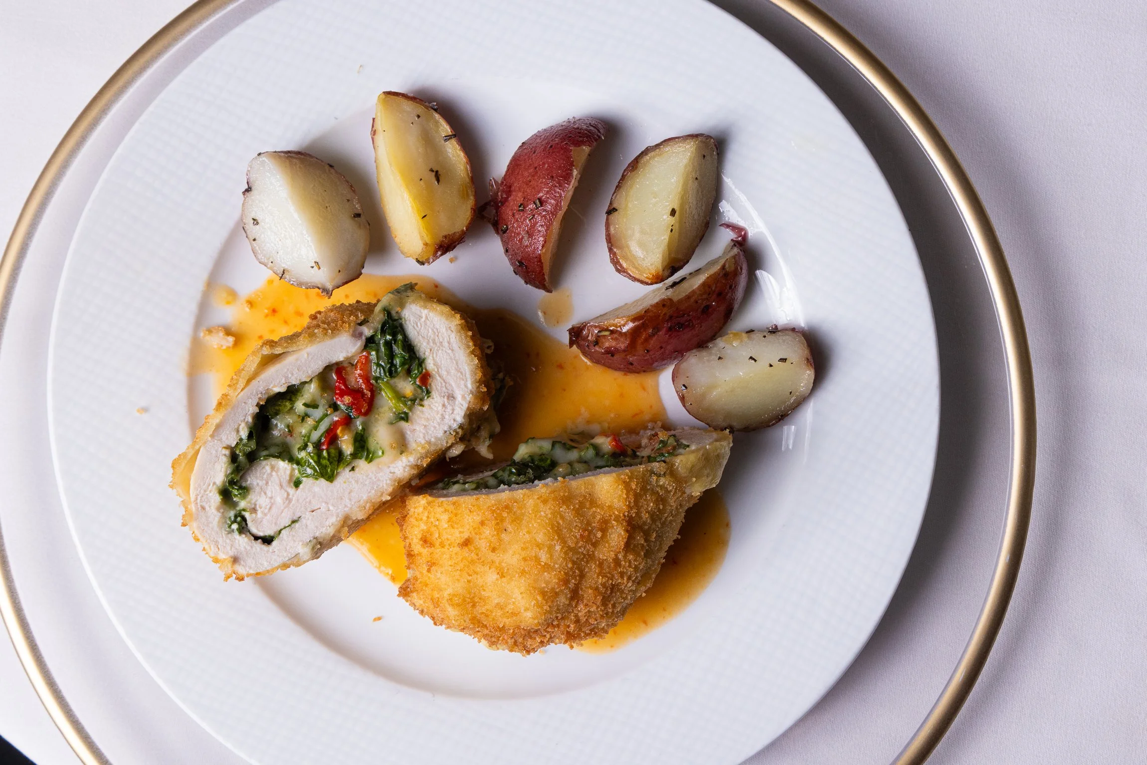 Breaded chicken stuffed with greens, red peppers, and herbs, served with roasted fingerling potatoes on a white plate with a gold rim.