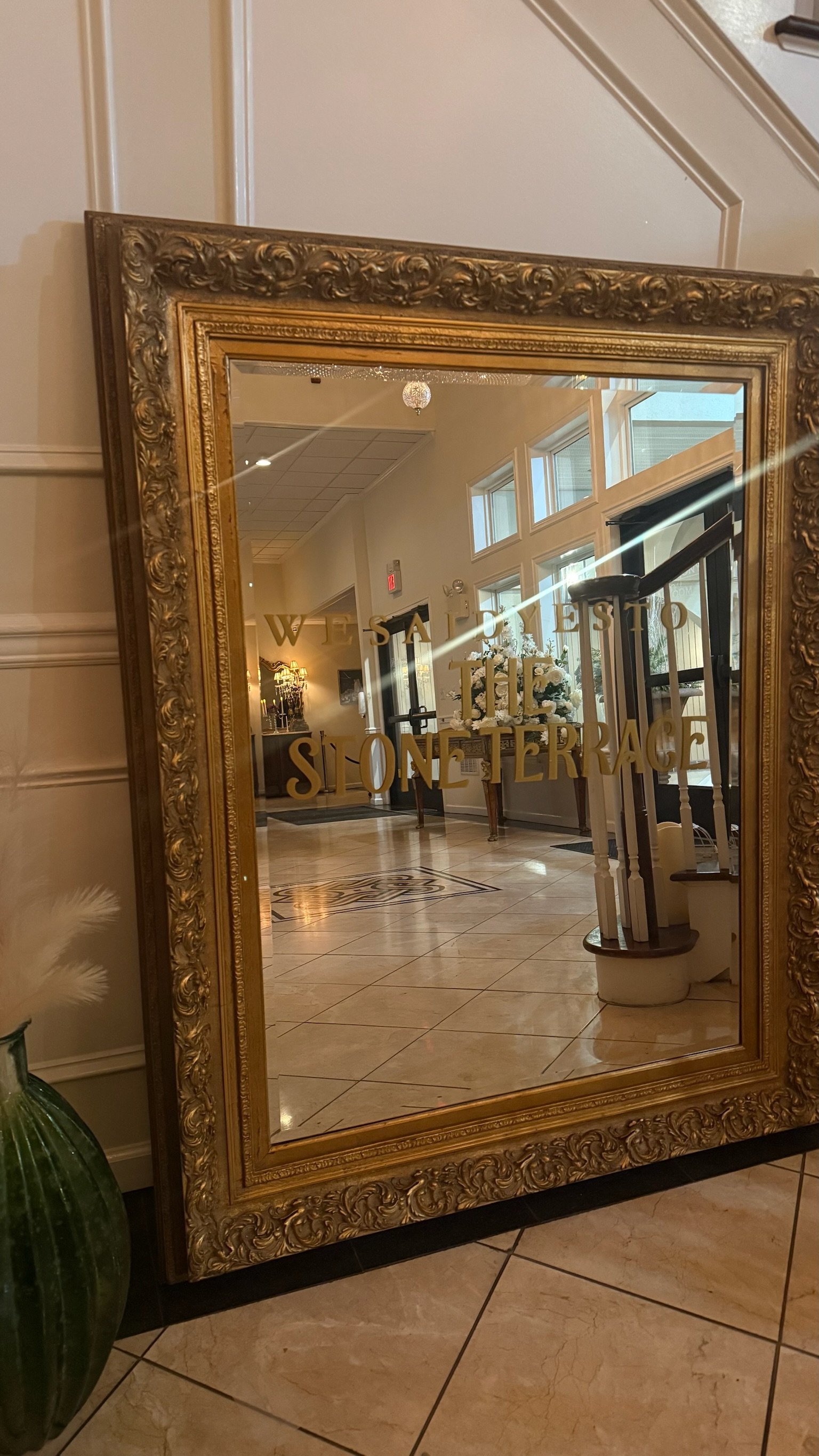 A large ornate gold-framed mirror in a hotel lobby. The mirror has gold lettering that reads "Welcome to the Stone Terrace." The lobby features a wooden console table with a floral arrangement, large windows, and a spiral staircase.