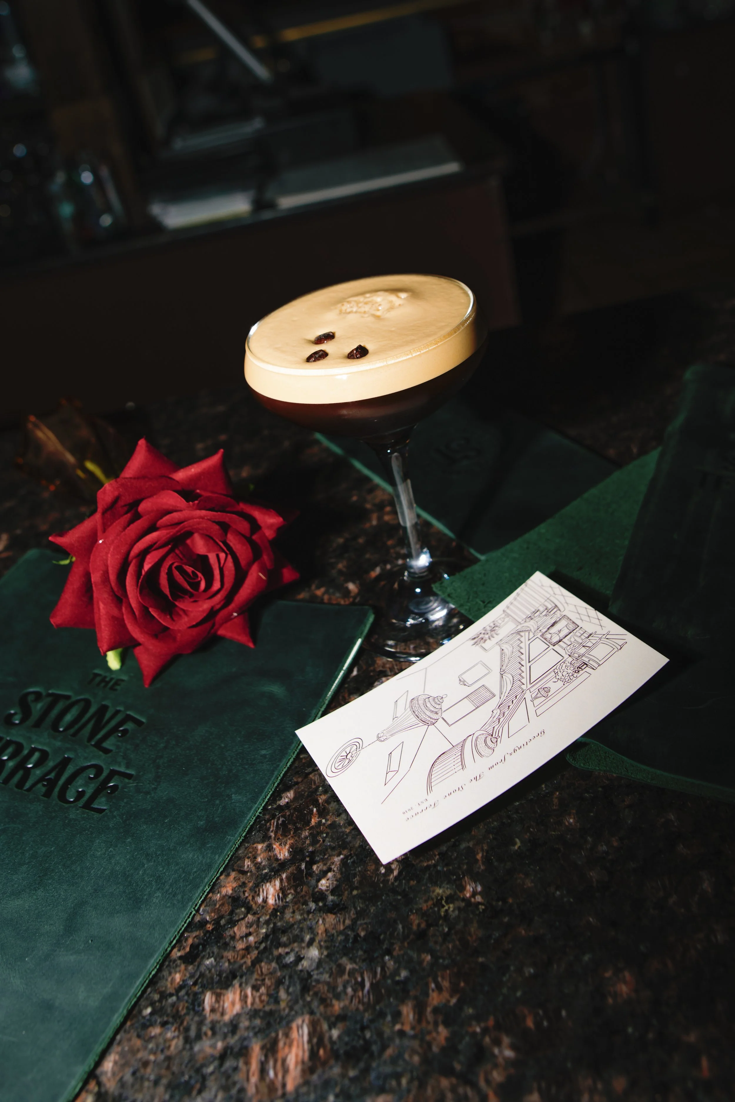A cocktail drink topped with coffee beans, a red rose, a green menu titled 'The Stone Garage,' and a sketch of a bridge on a napkin on a dark granite bar counter.