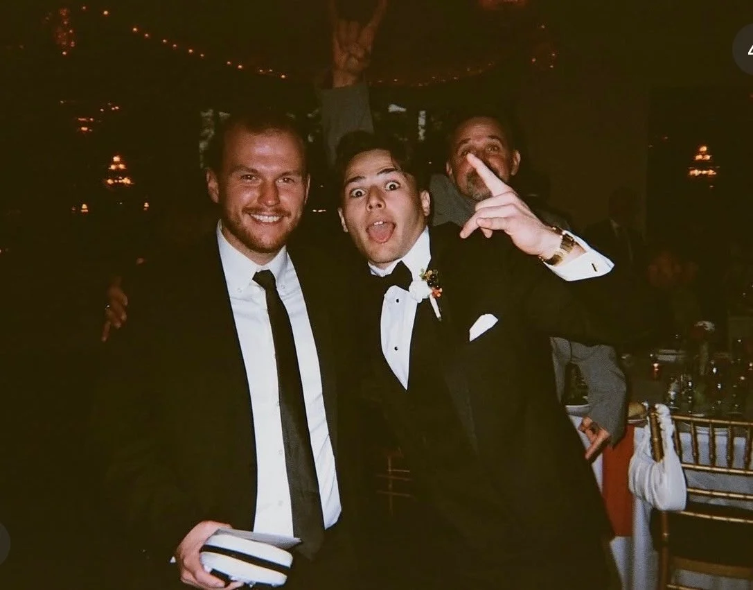 Three men in formal attire at a festive event, with one making a gesture and all smiling.