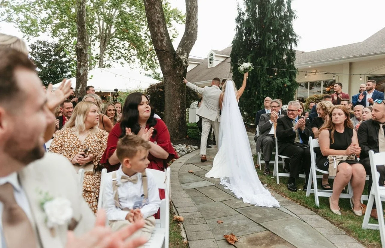 Bride and groom walking down the aisle during an outdoor wedding ceremony, surrounded by seated guests clapping and watching, with trees and a house in the background.