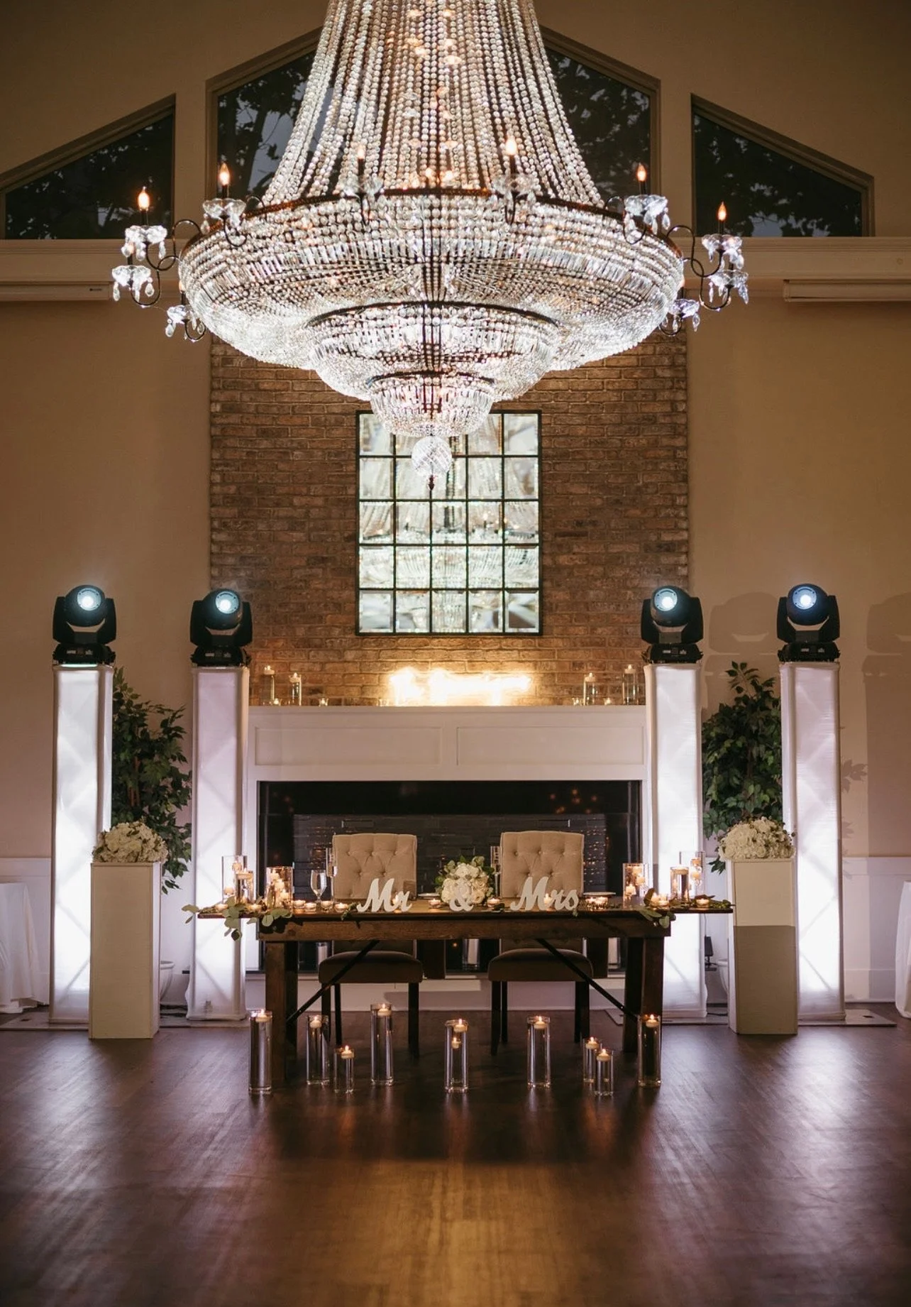 Elegant wedding reception area with a large chandelier hanging from the ceiling, a brick fireplace, a window above it, and a table with 'Mr & Mrs' signs, floral arrangements, candles, and decorative lighting in a spacious hall.