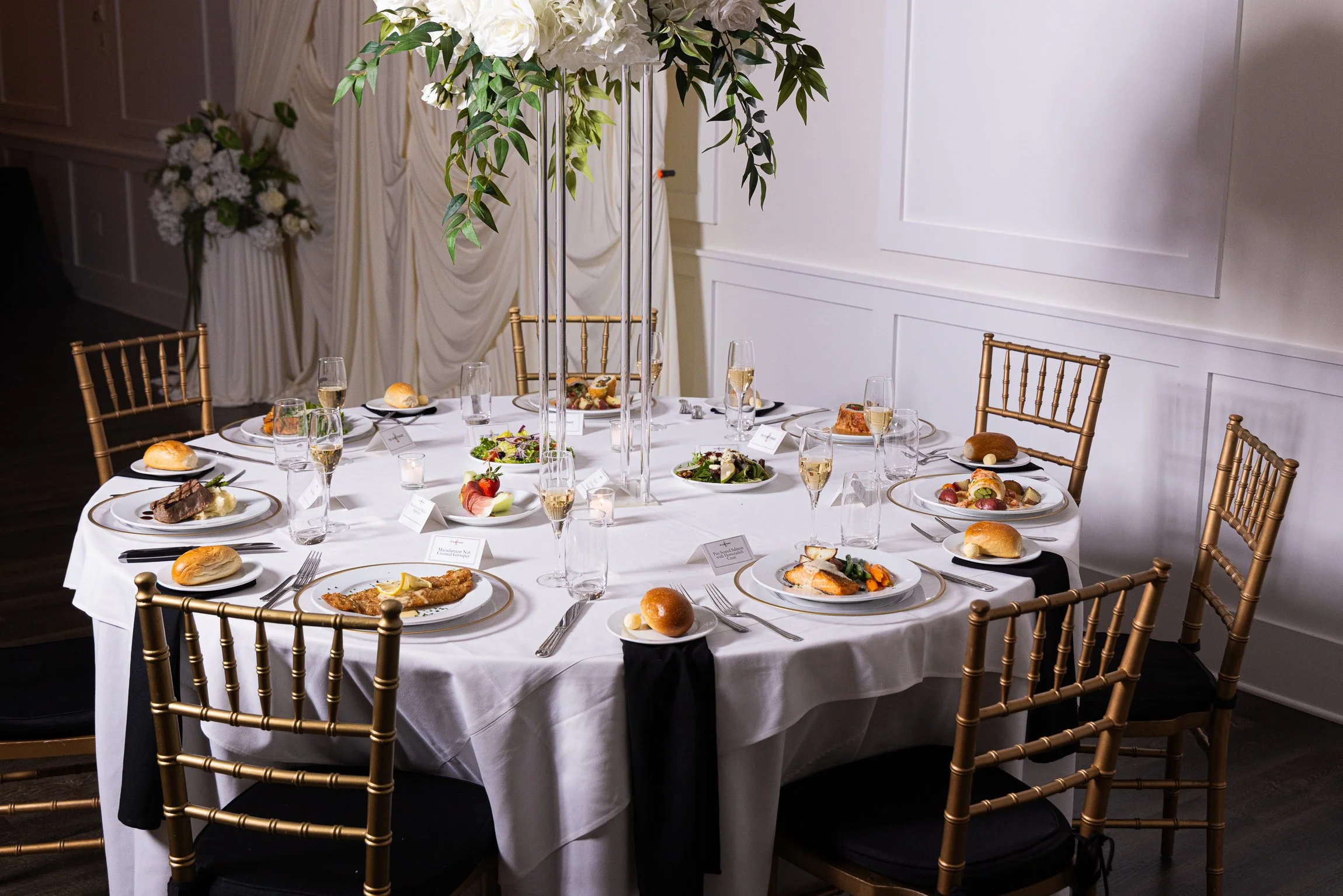 Round banquet table set for a formal event, with food plates, champagne glasses, and a tall floral centerpiece, surrounded by gold Chiavari chairs, a white tablecloth, and elegant decorations.