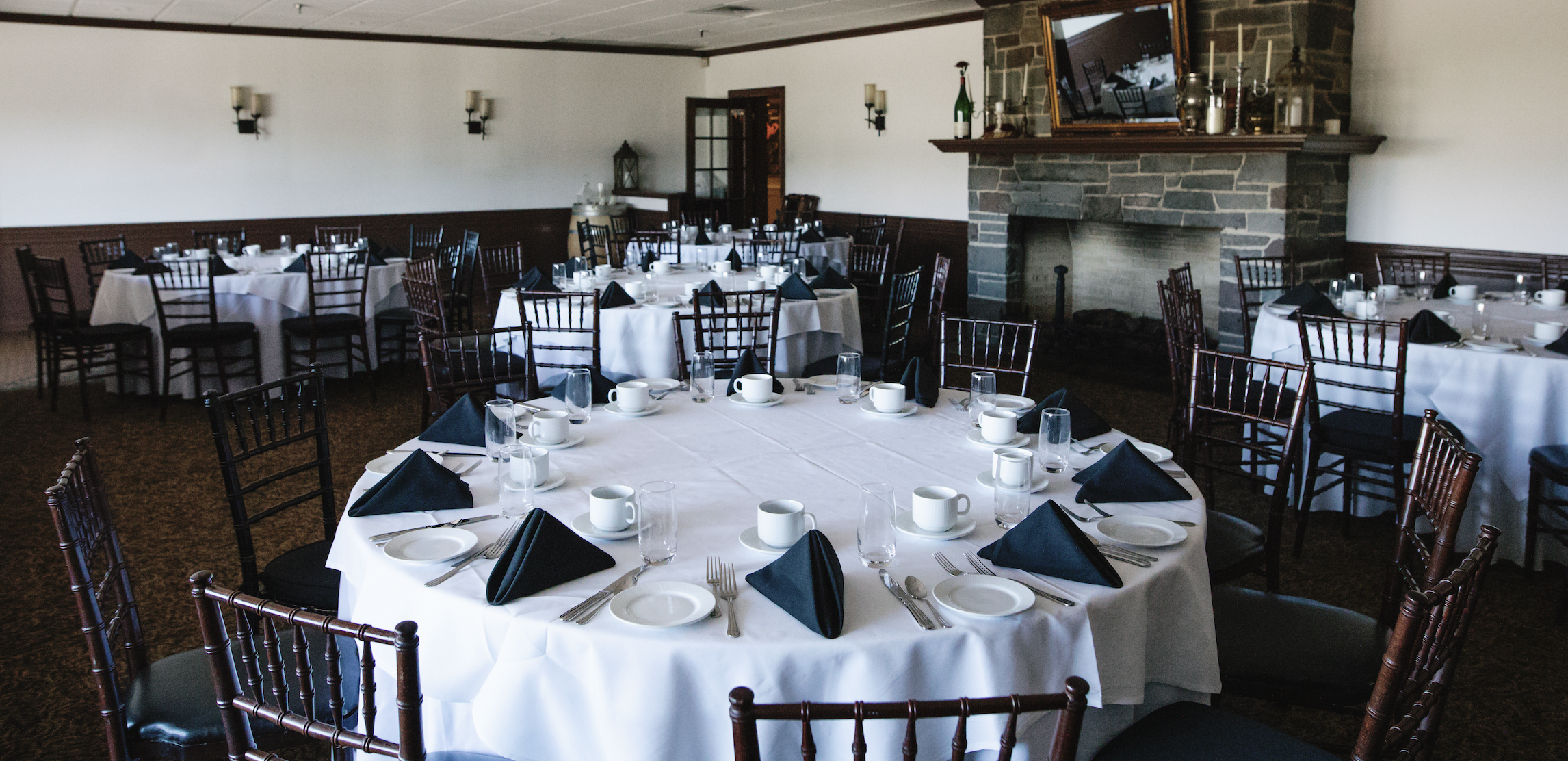 A restaurant dining room with multiple round tables covered in white tablecloths. Each table is set with black napkins, white plates, silverware, water glasses, and coffee cups. The room has dark wooden chairs with black cushions, a stone fireplace with a mirror above it, and a wall-mounted TV. The setting appears ready for a formal event or gathering.