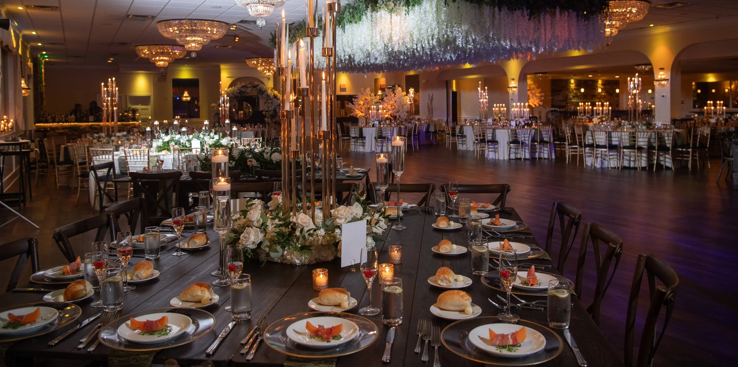 Elegant banquet hall decorated for an event with round tables and a long rectangular table, floral centerpieces, candles, chandeliers, and ambient lighting.