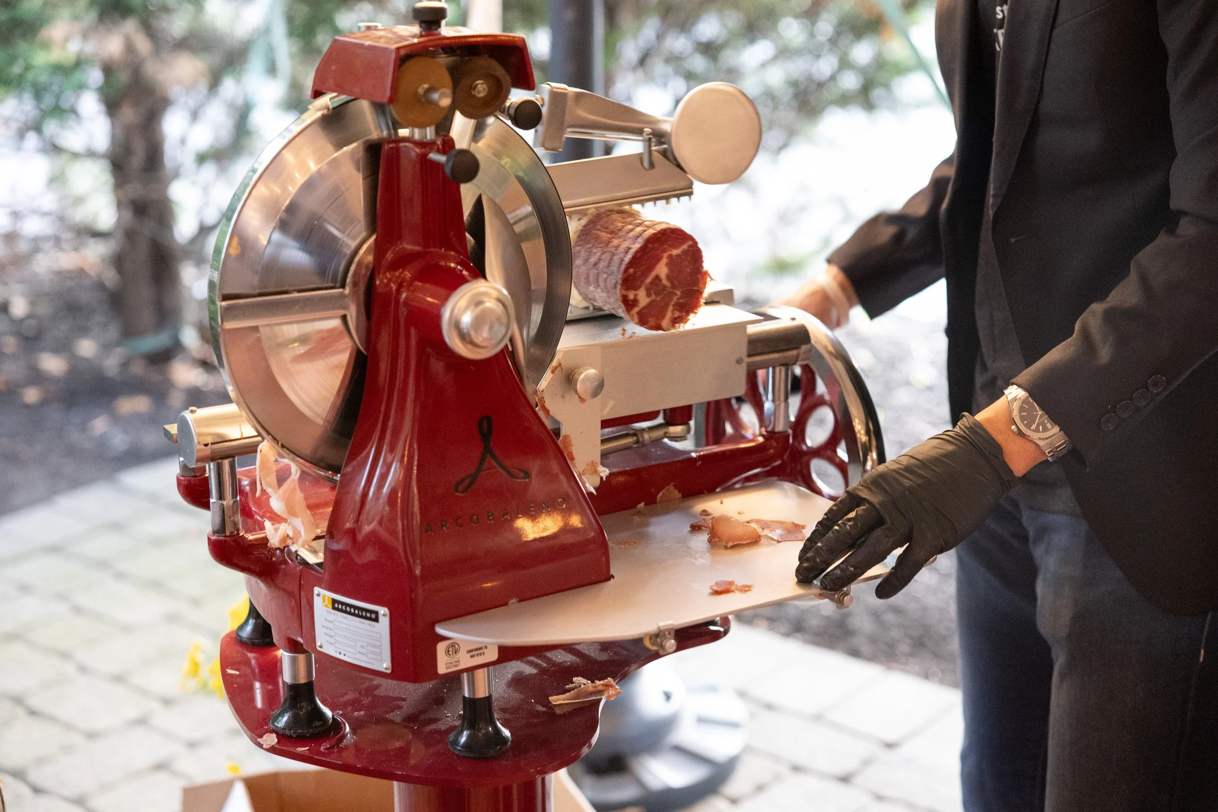 A person slicing prosciutto from a cured ham on a vintage red meat slicer outdoors.