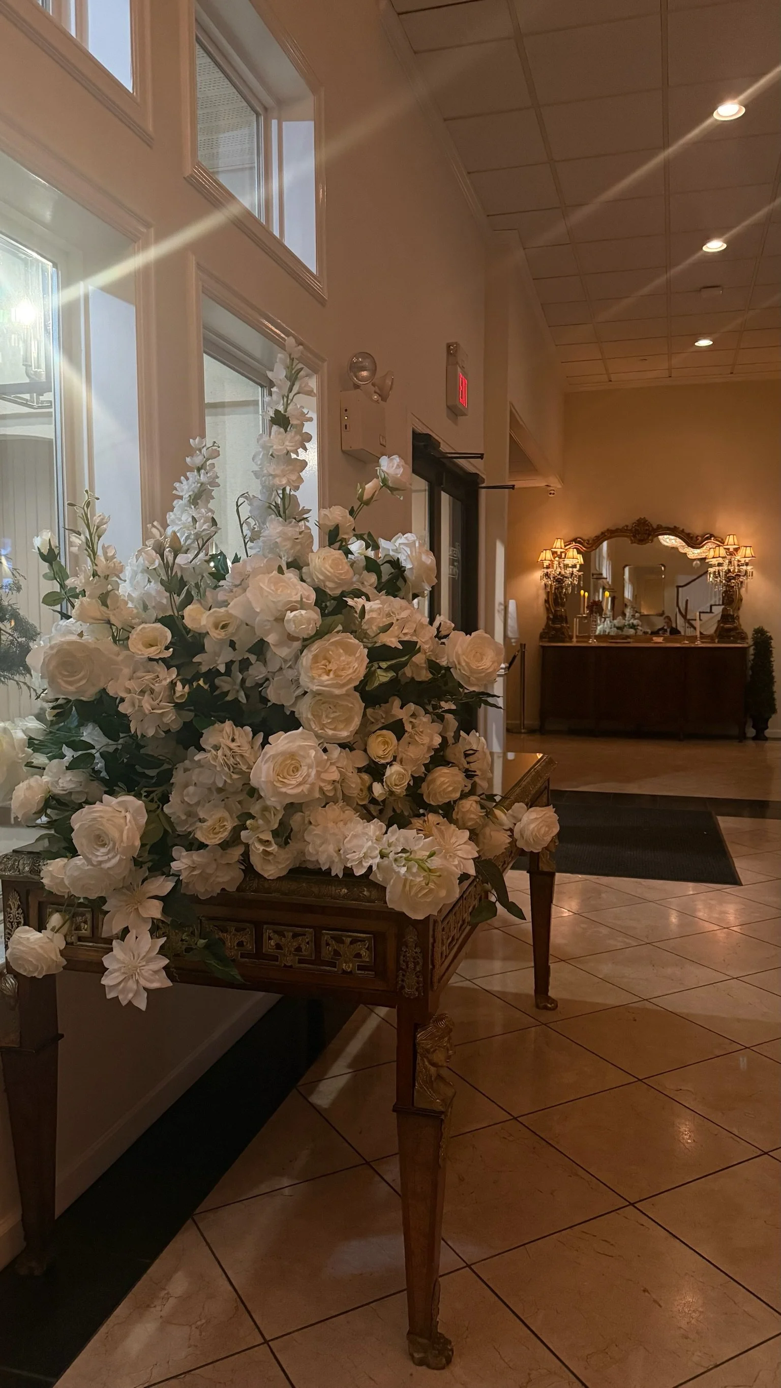 A large white floral arrangement with roses and other flowers on a decorative wooden table in the lobby of a hotel or event venue, with a mirror and warm lighting in the background.