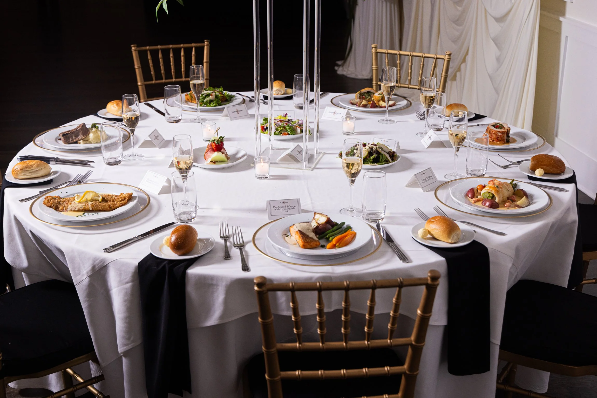 A round banquet table set for a formal dinner with a white tablecloth, multiple plates of appetizers and entrees, wine glasses filled with white wine, small candles, and decorated with elegant black napkins.