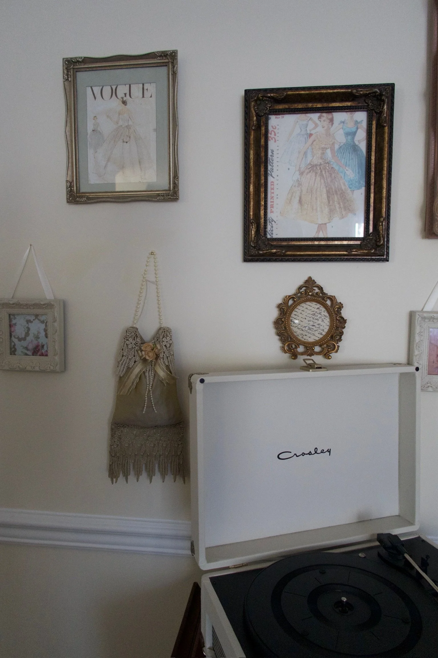 A vintage-style wall with framed fashion illustrations, ornate decorative mirrors, and a lace wall hanging, alongside a Crosley record player.