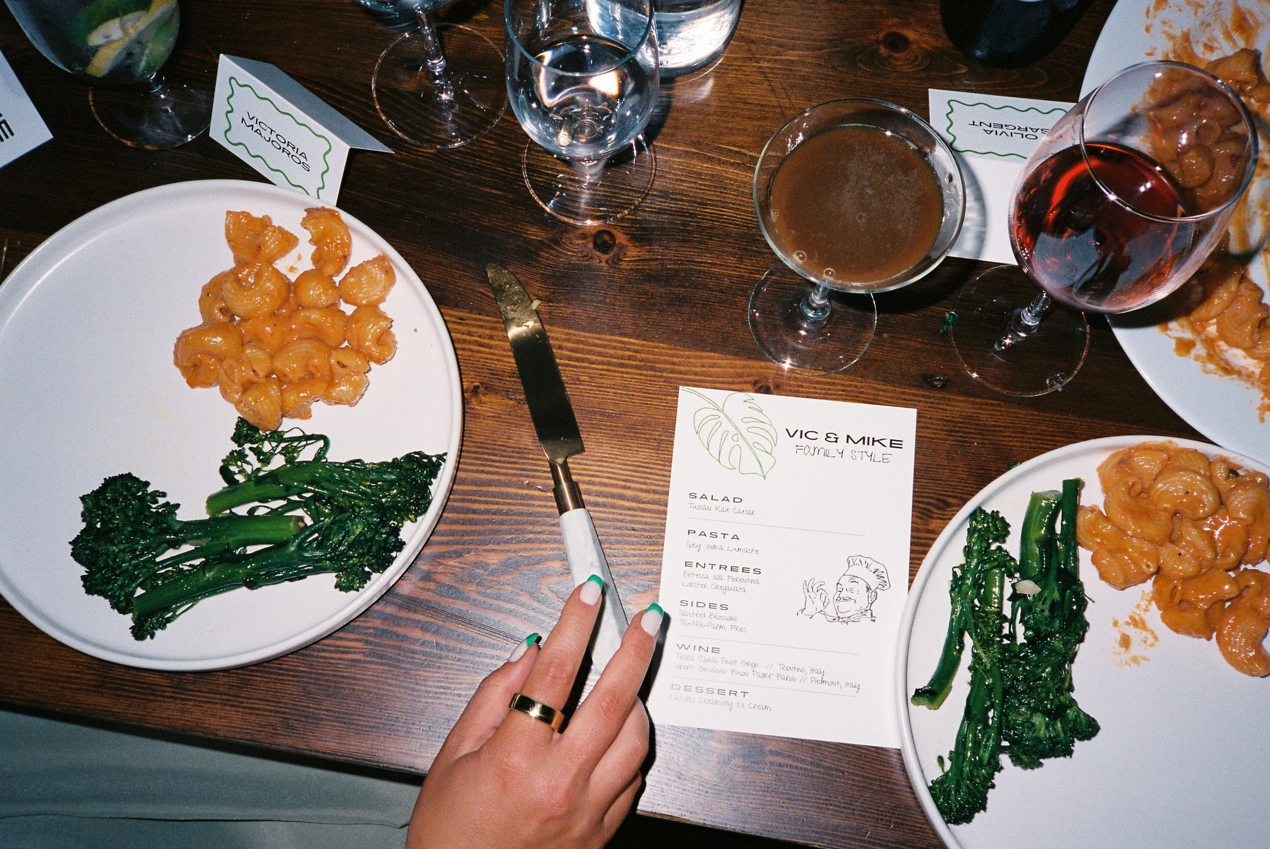 A table setting at a restaurant with two white plates of food, two glasses of wine, a glass of beer, a knife, a menu, and a hand with painted nails and a ring.