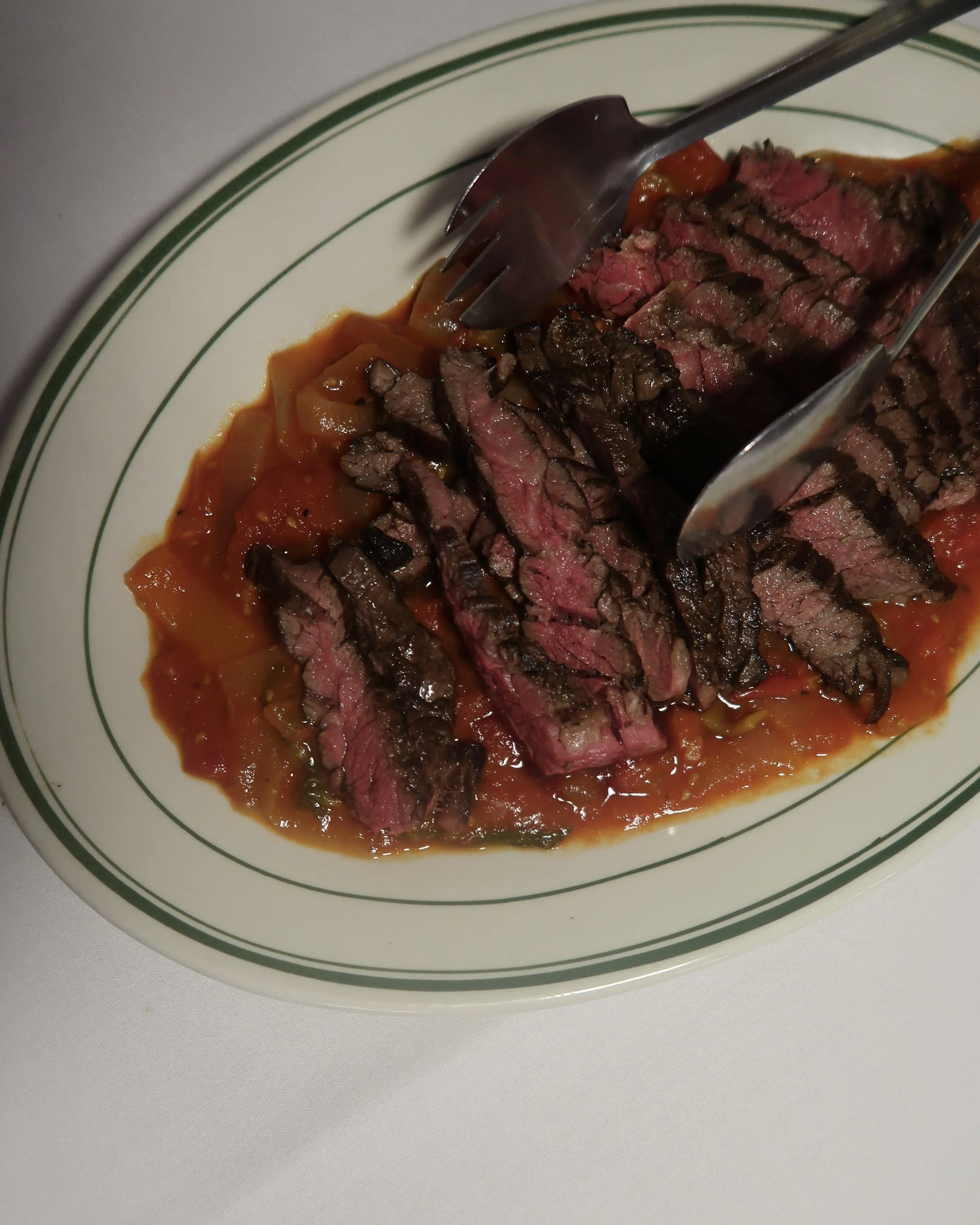 Slices of medium-rare grilled steak placed on a bed of tomato-based sauce on a white plate with green rim.