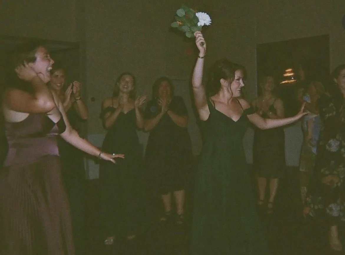 Women celebrating at a party, with one woman holding a bouquet of flowers in the air and the others clapping and smiling.