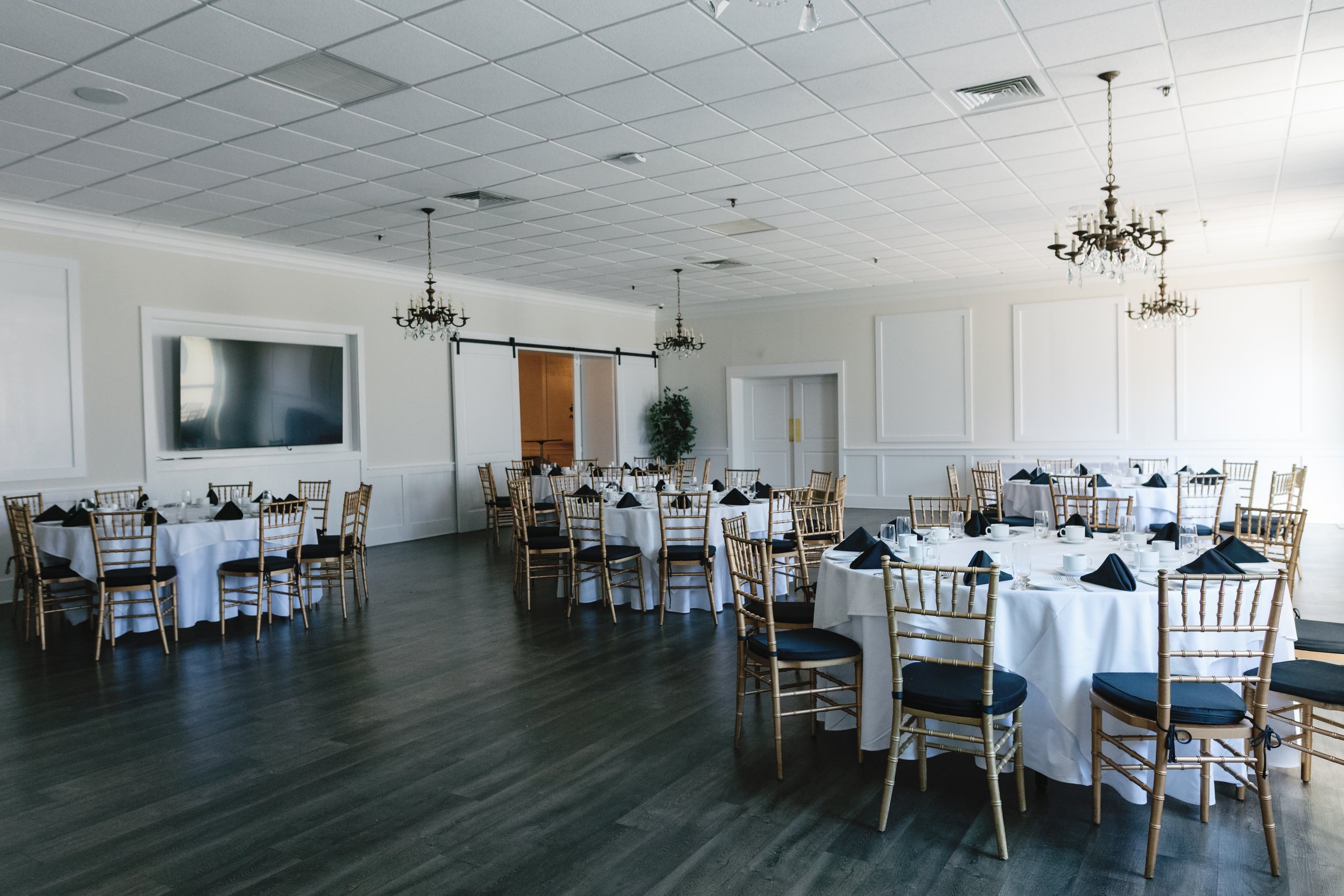 A large, elegant banquet hall with white walls, dark wooden floors, and columns. Several round tables are set with white tablecloths, black napkins, and glassware. Gold-colored chairs with black cushions surround the tables. Chandeliers hang from the ceiling, and a large flat-screen TV is mounted on the wall. There are also decorative wall moldings and a sliding barn door in the background.