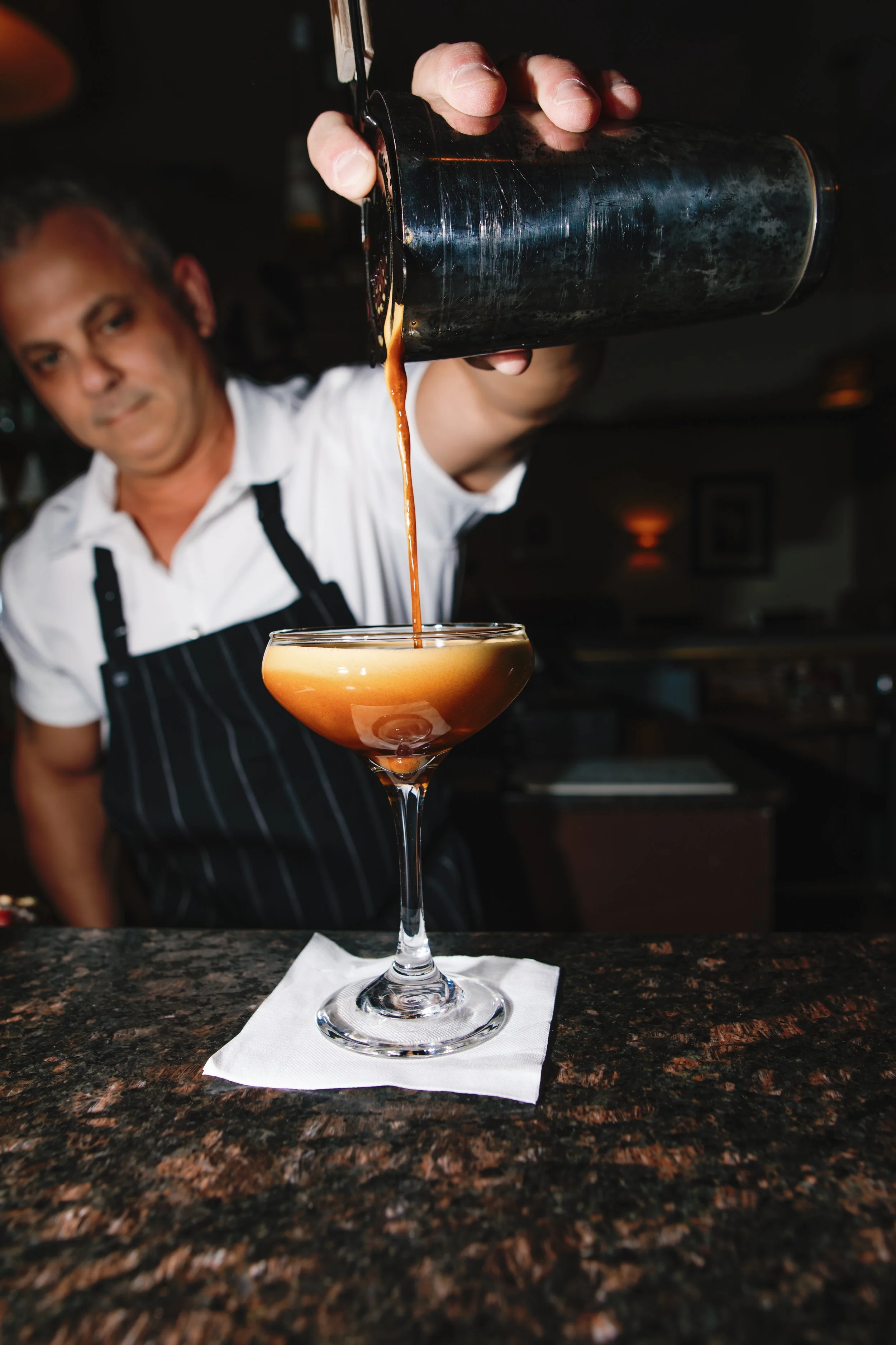 Person in a white shirt and black apron pouring cocktail from a shaker into a coupe glass, which is placed on a white napkin on a dark countertop.