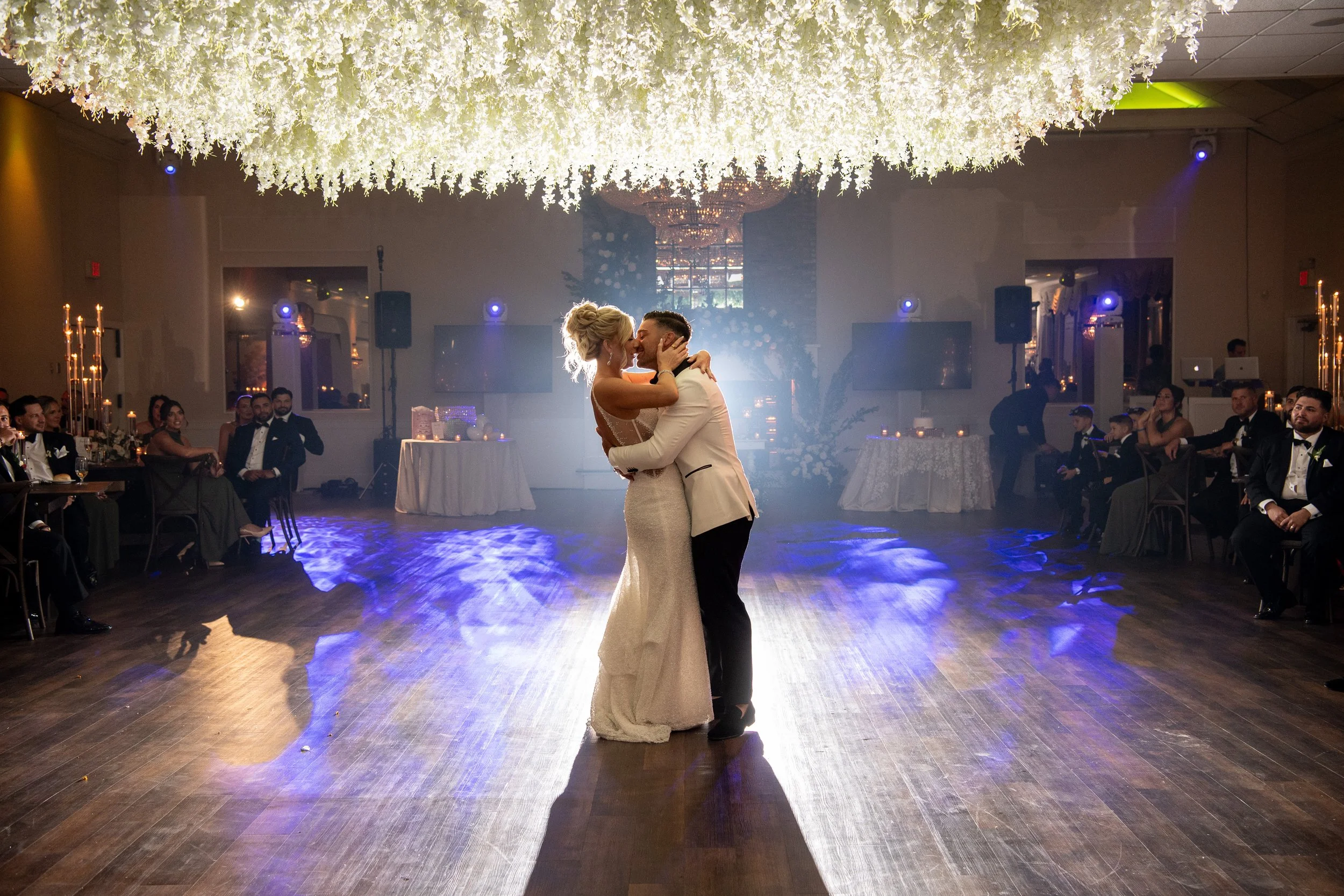 A bride and groom dance together at their wedding reception, illuminated by a spotlight, with guests seated around the dance floor and floral decorations overhead.