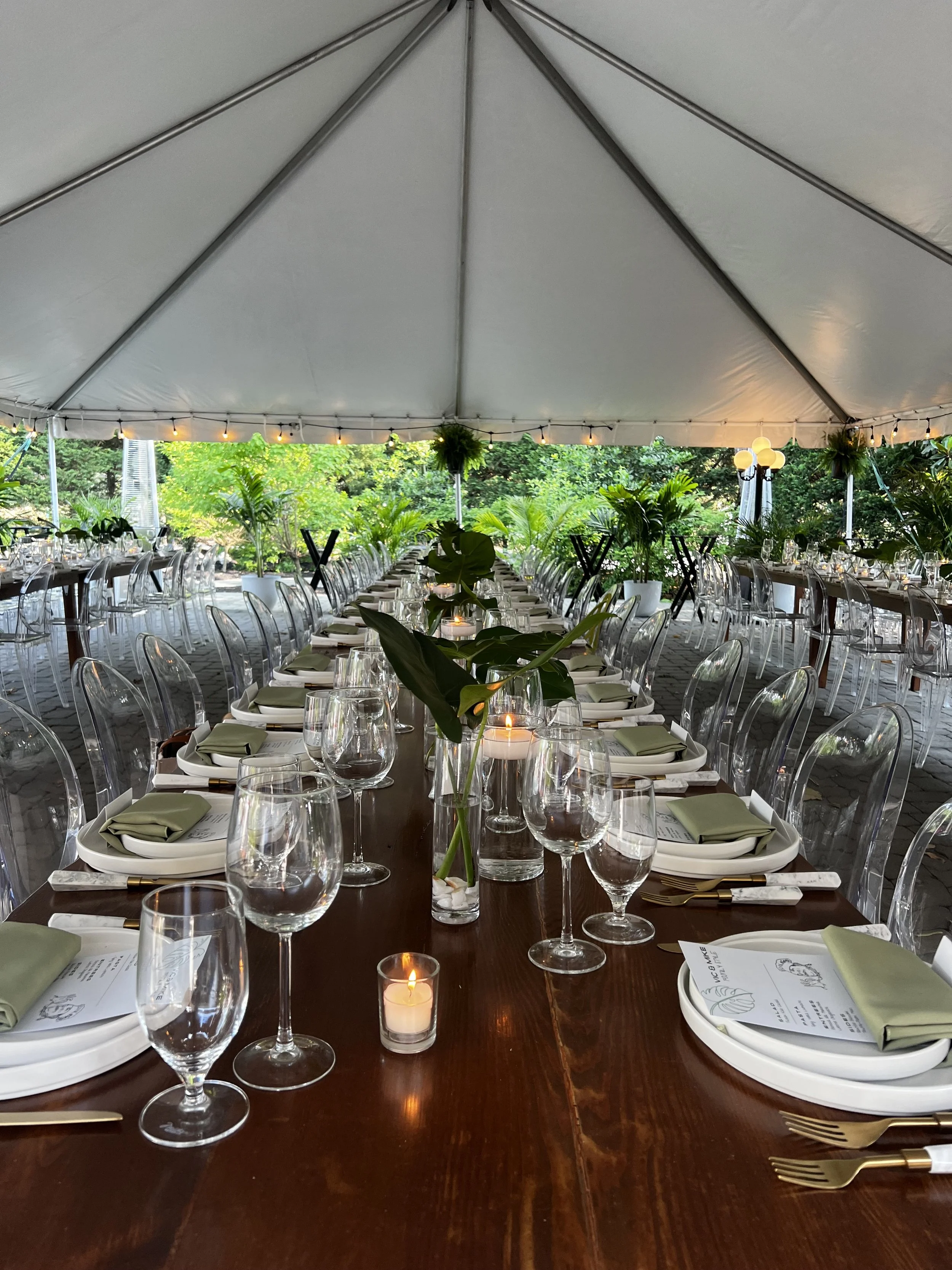 Elegant outdoor dining setup under a large white tent with long wooden table, clear chairs, green napkins, glassware, candles, and lush greenery in the background.