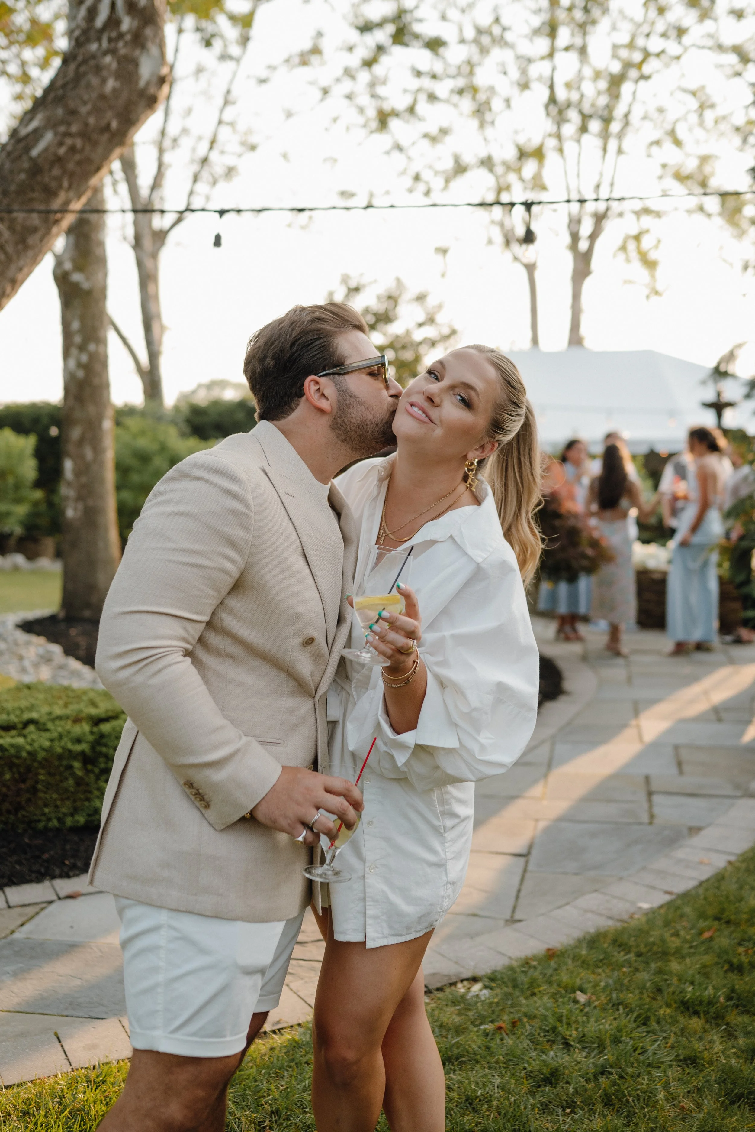 A man kisses a woman on the cheek at an outdoor gathering, both holding cocktails, with other guests in the background and trees overhead.