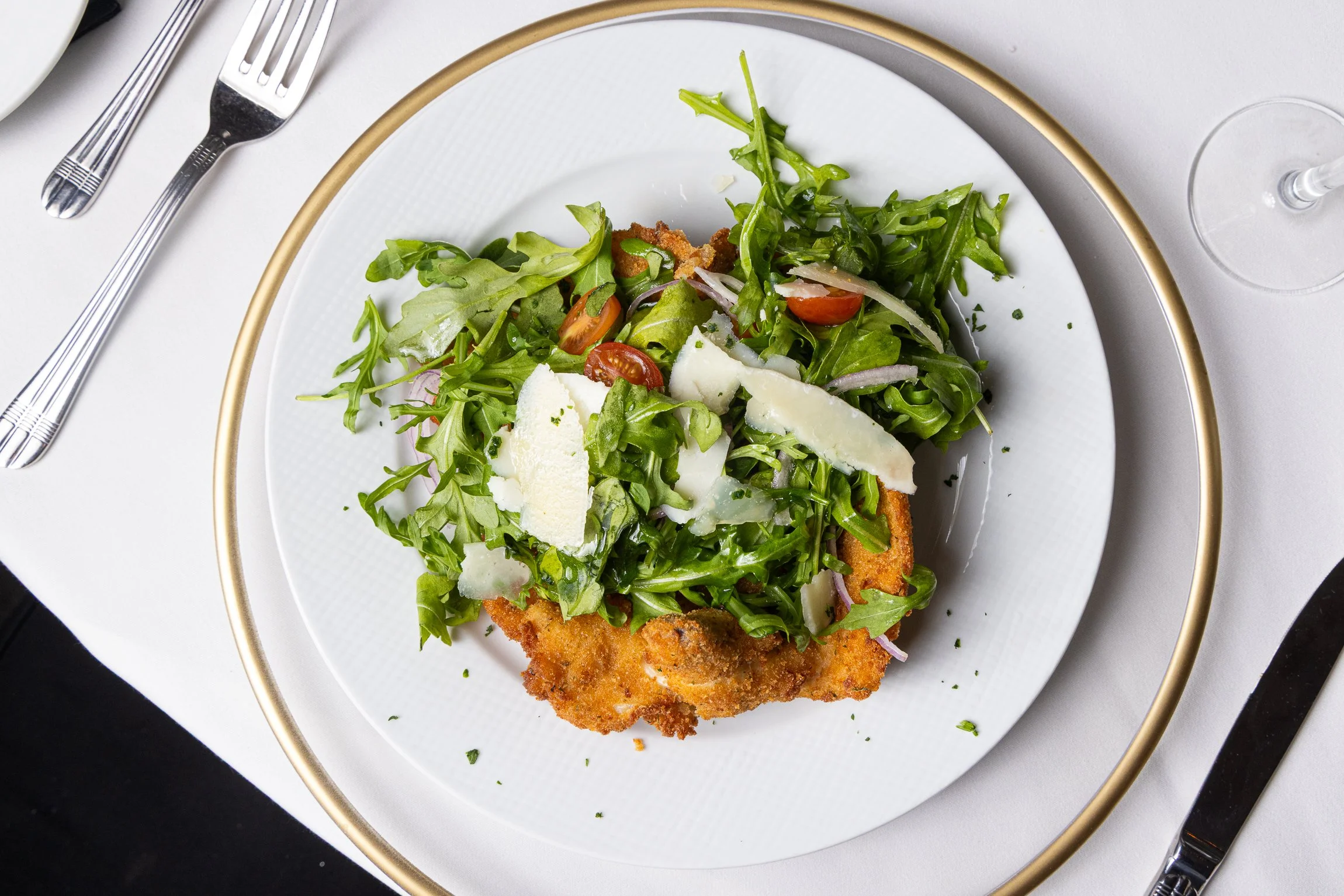 Salad with mixed greens, cherry tomatoes, onions, and shaved cheese on top of a breaded and fried piece of chicken or fish on a white plate with a gold rim, set on a white tablecloth with silverware.