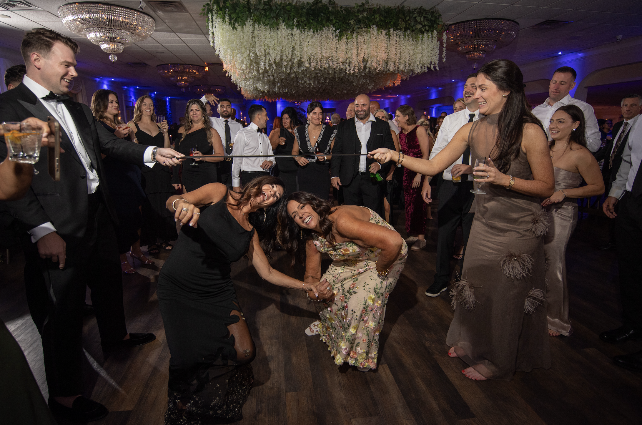 A group of people dancing and celebrating at a party, with some holding drinks, in a decorated ballroom with hanging floral arrangements and chandeliers.