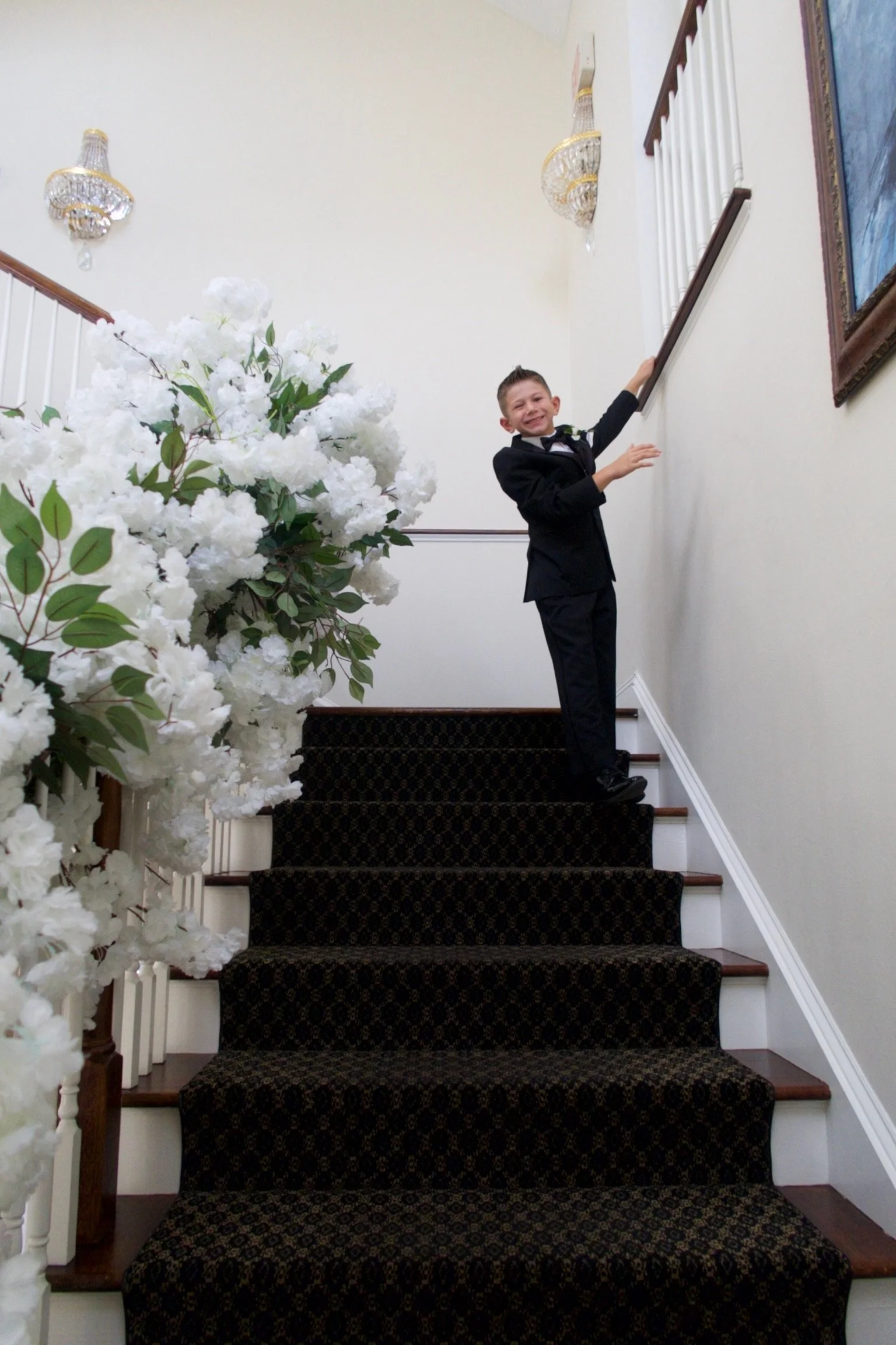 A young boy in a tuxedo standing on a staircase, smiling and holding onto the handrail.