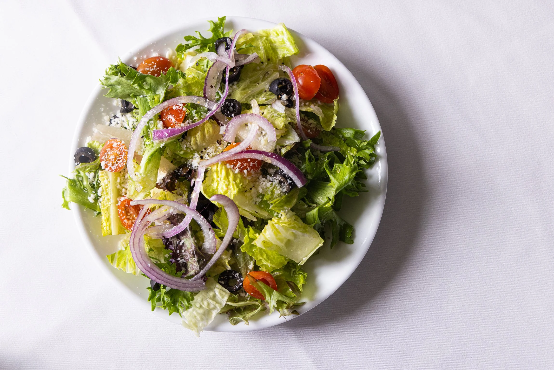 A bowl of mixed salad with lettuce, cherry tomatoes, black olives, red onion slices, and shredded cheese on a white background.
