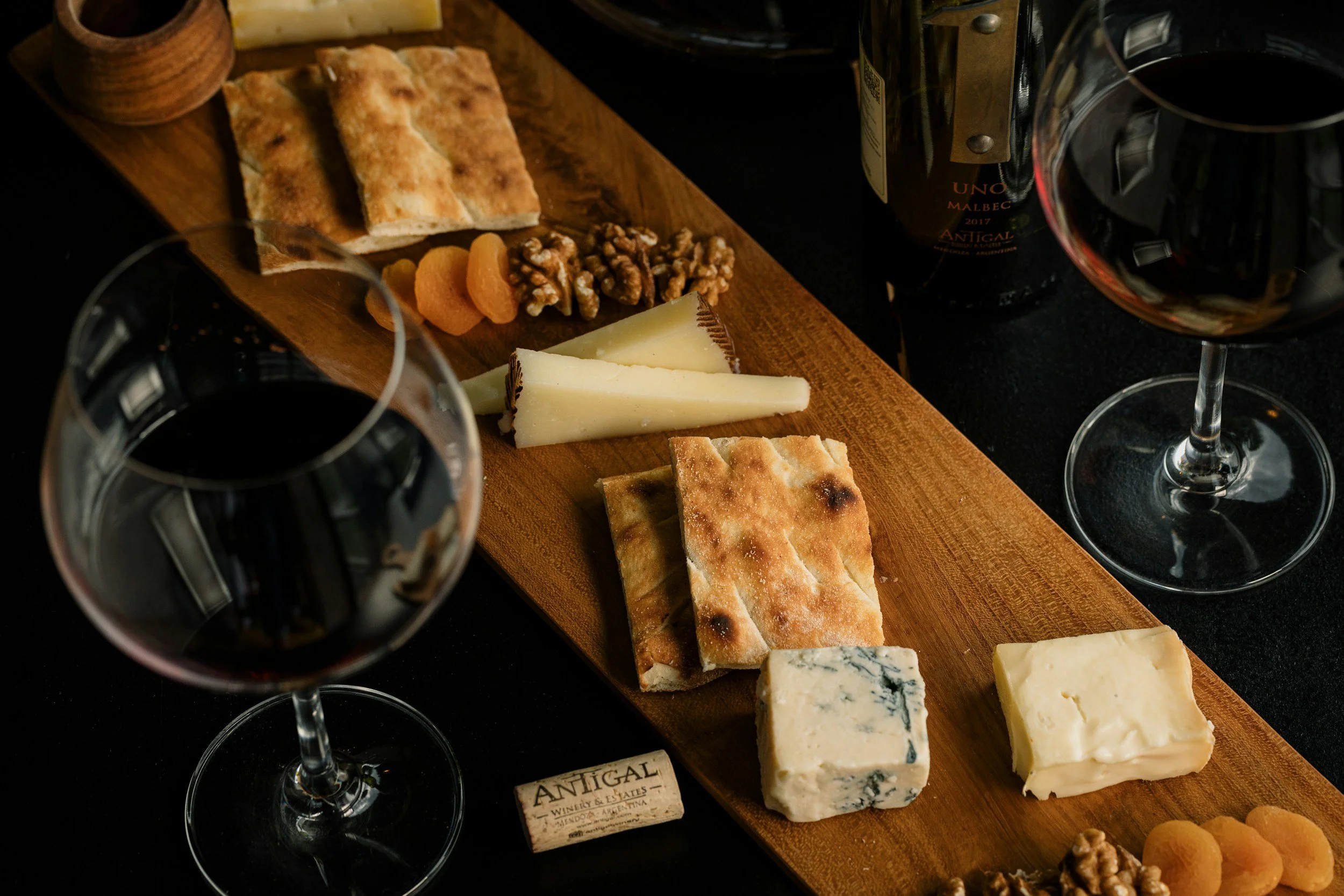 A cheese and charcuterie board with various cheeses, dried apricots, walnuts, flatbread, and preserved fruits, accompanied by two glasses of red wine.
