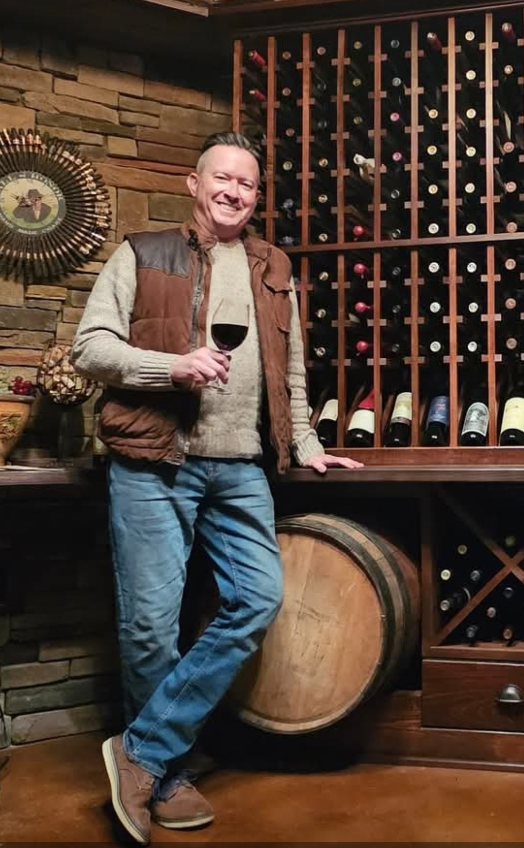 A man holding a glass of red wine, standing in a wine cellar with a wall of wine bottles behind him and a large wine barrel underneath the counter.
