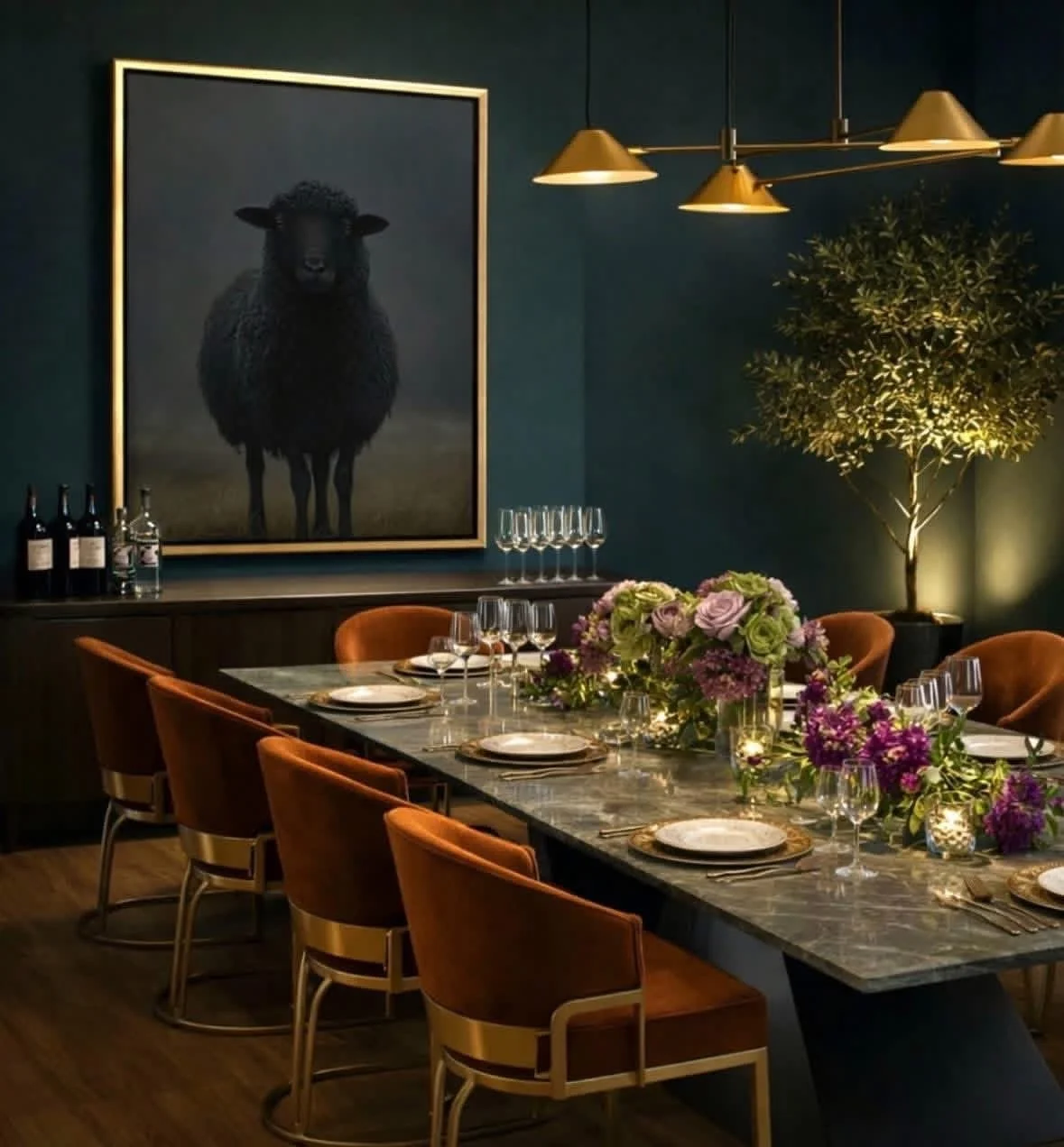 A elegantly set dining table with plates, wine glasses, and floral centerpieces in a stylish dining room, decorated with a large framed painting of a black sheep, a potted tree illuminated by a floor lamp, and modern gold ceiling lights.