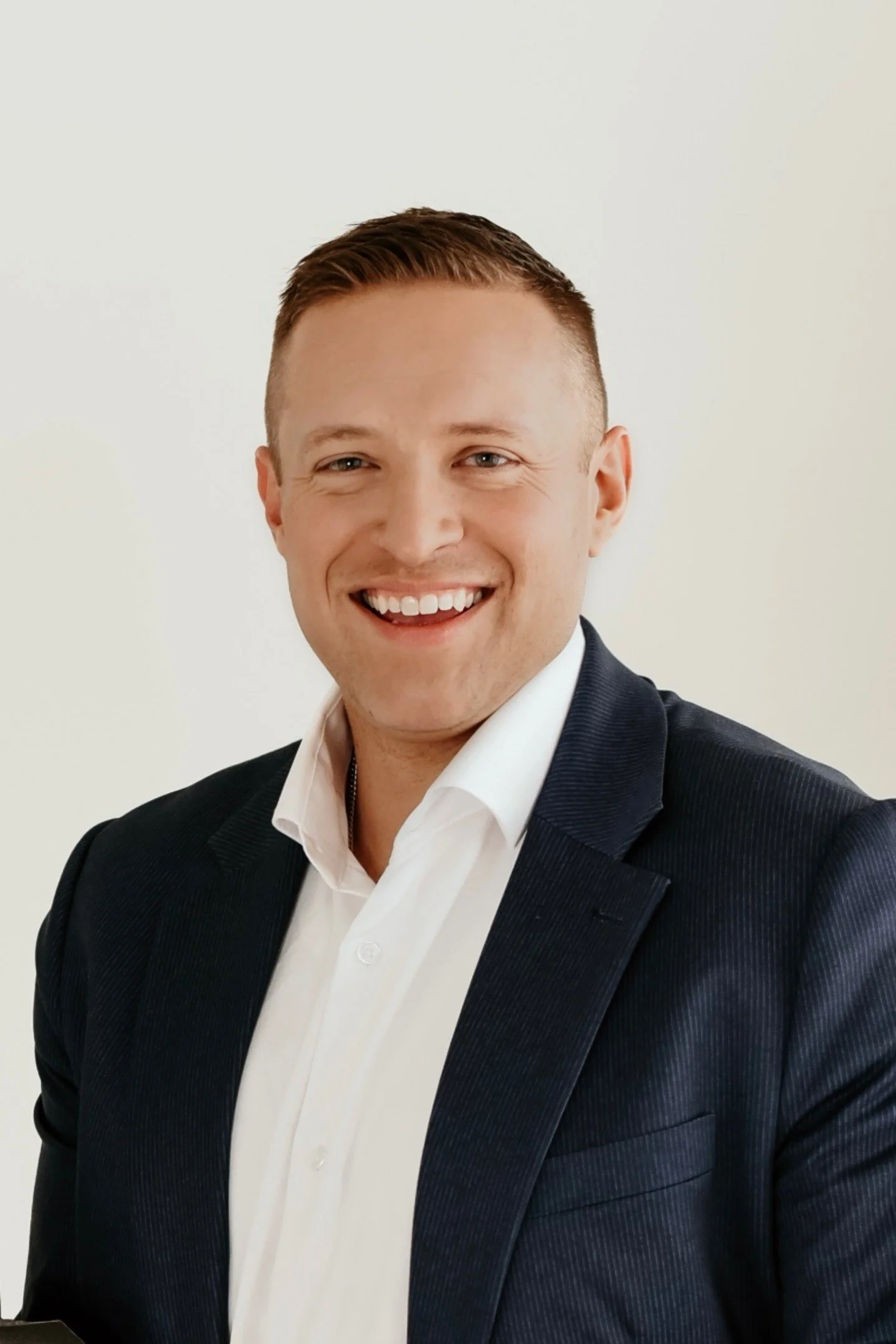 A young man smiling, wearing a dark suit jacket and white shirt, posed against a plain light background.