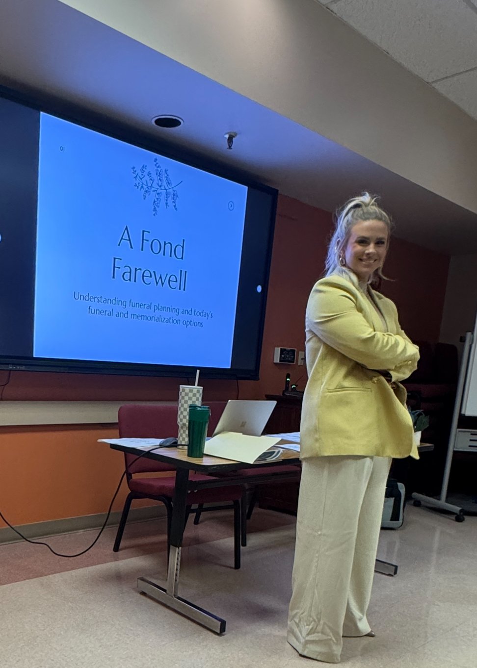 A woman standing with her arms crossed in front of a presentation screen that reads 'A Fond Farewell' and discusses funeral planning options. She is in a conference room with a table, two drinks, a laptop, and papers.