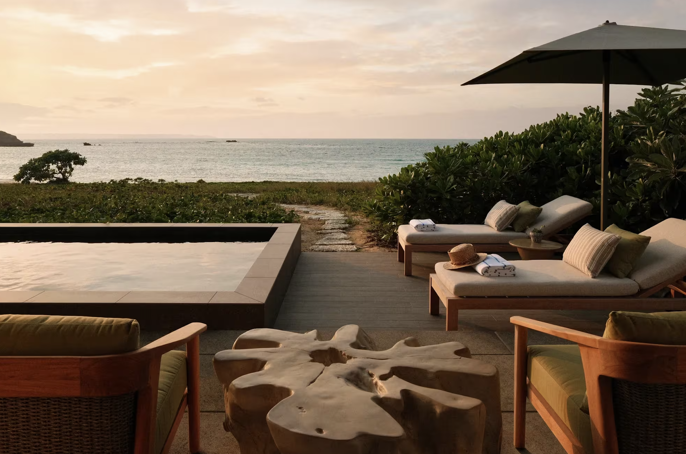 Sunset view of a beachside resort with a hot tub, lounge chairs with pillows, a straw hat, towels, an umbrella, and a wooden table on a deck overlooking the ocean with greenery.