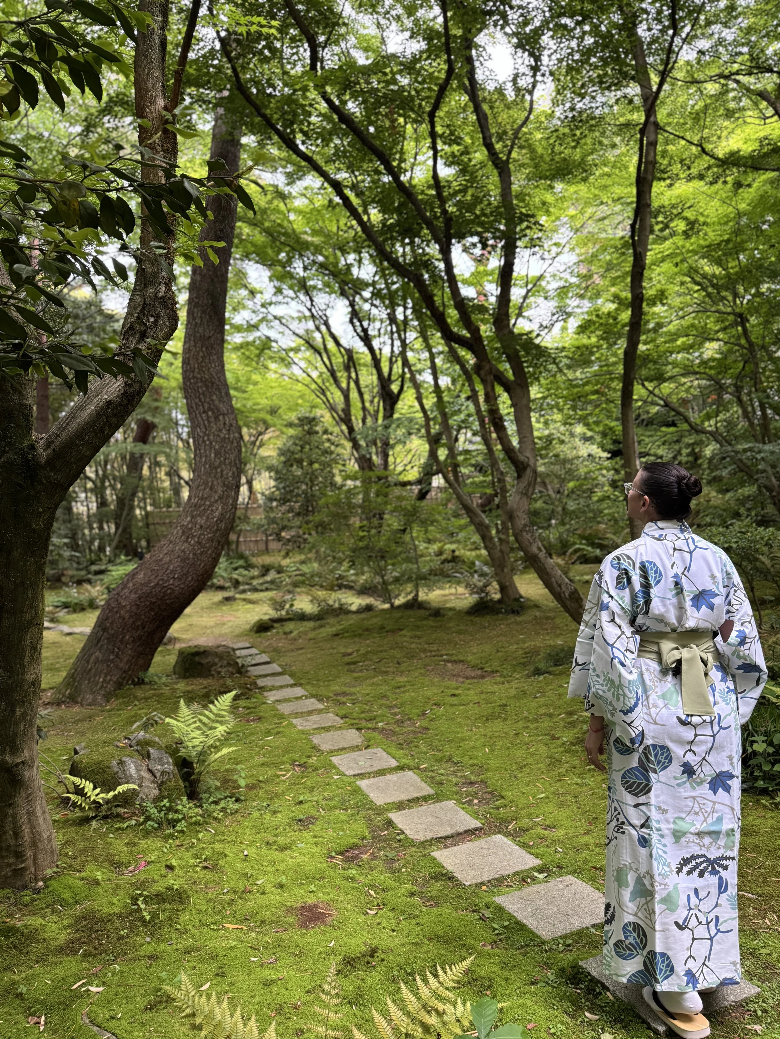 A person wearing a white yukata with blue and green leaf patterns walking through a lush Japanese-style garden with trees, ferns, and stepping stones.