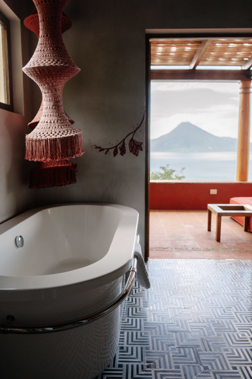 A white oval bathtub beside a dark gray wall with a painted flower and a large window overlooking a mountain and water. A red crocheted lamp hangs above the bathtub, and a towel is draped over a metal bar attached to the tub.