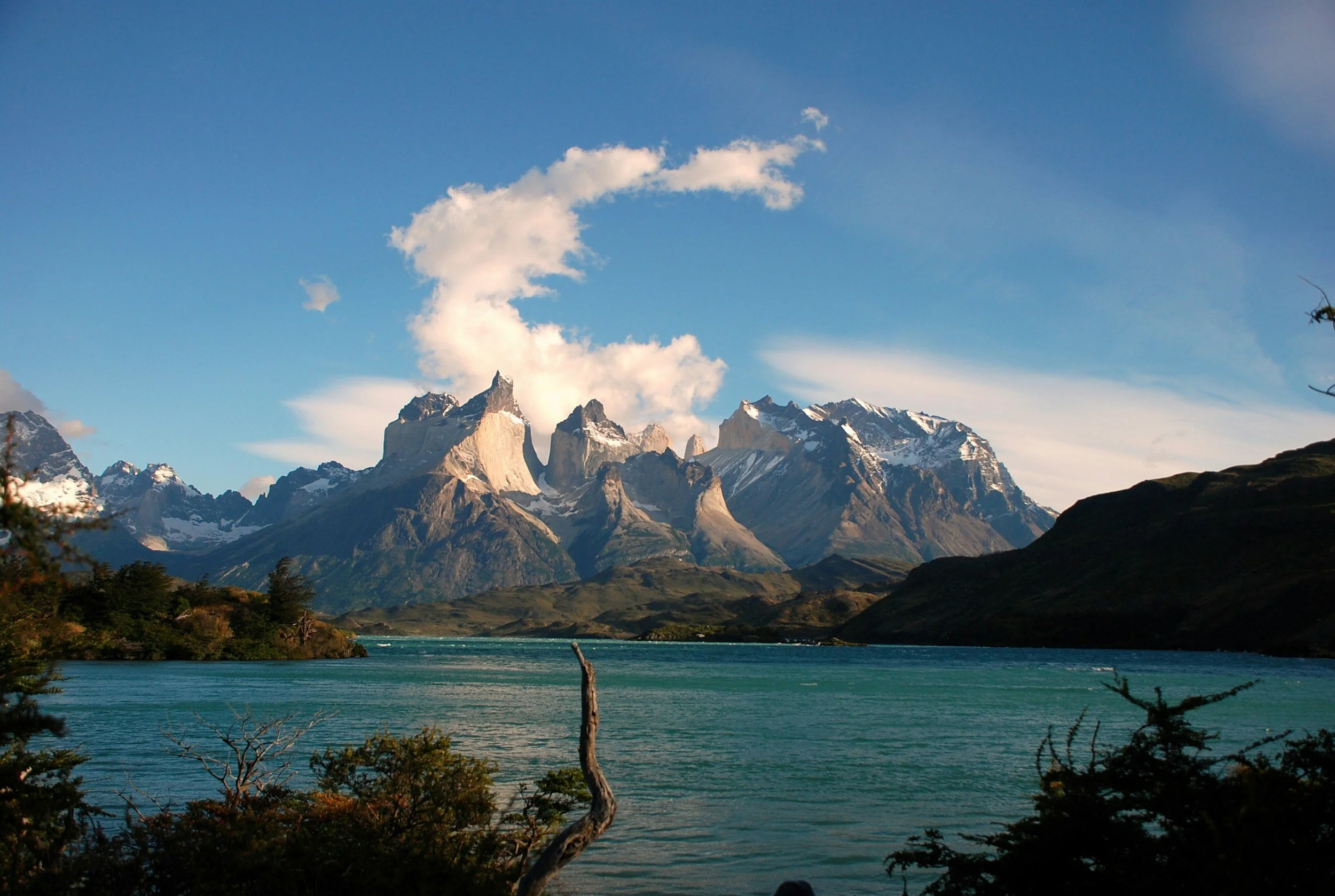 Scenic view of a mountain range with snow-capped peaks overlooking a lake with a few trees and bushes in the foreground under a blue sky with some clouds.