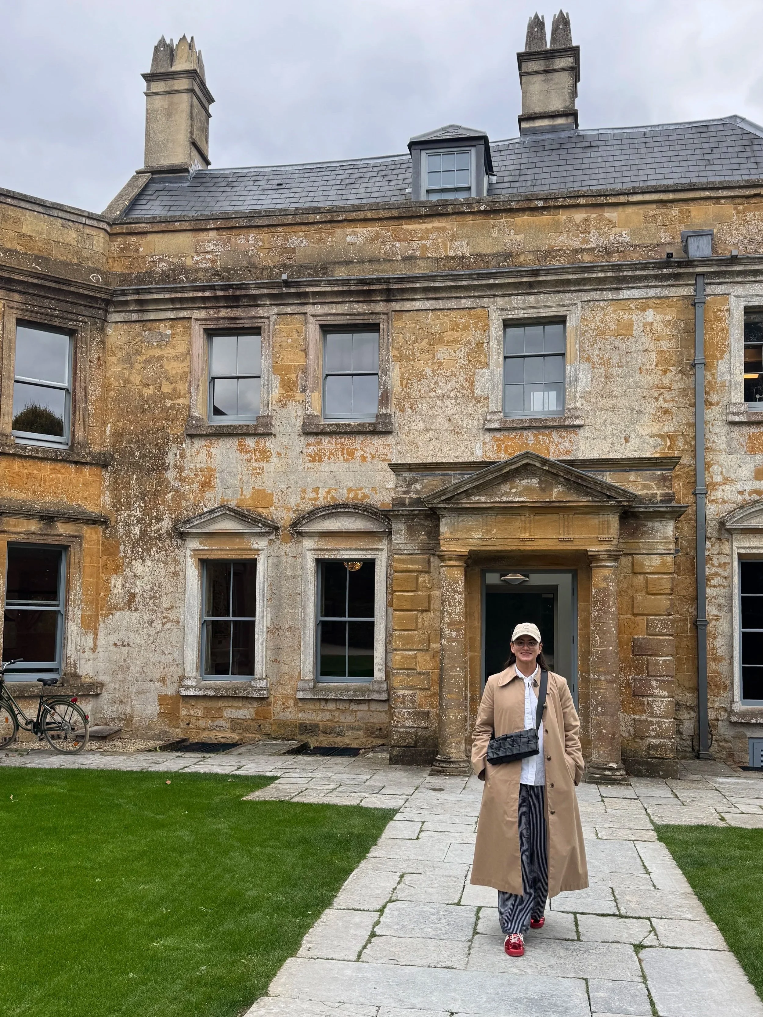 A woman wearing a beige trench coat, white shirt, gray striped pants, pink sneakers, and a white hat standing outside a historic stone building with multiple windows and a grassy lawn.