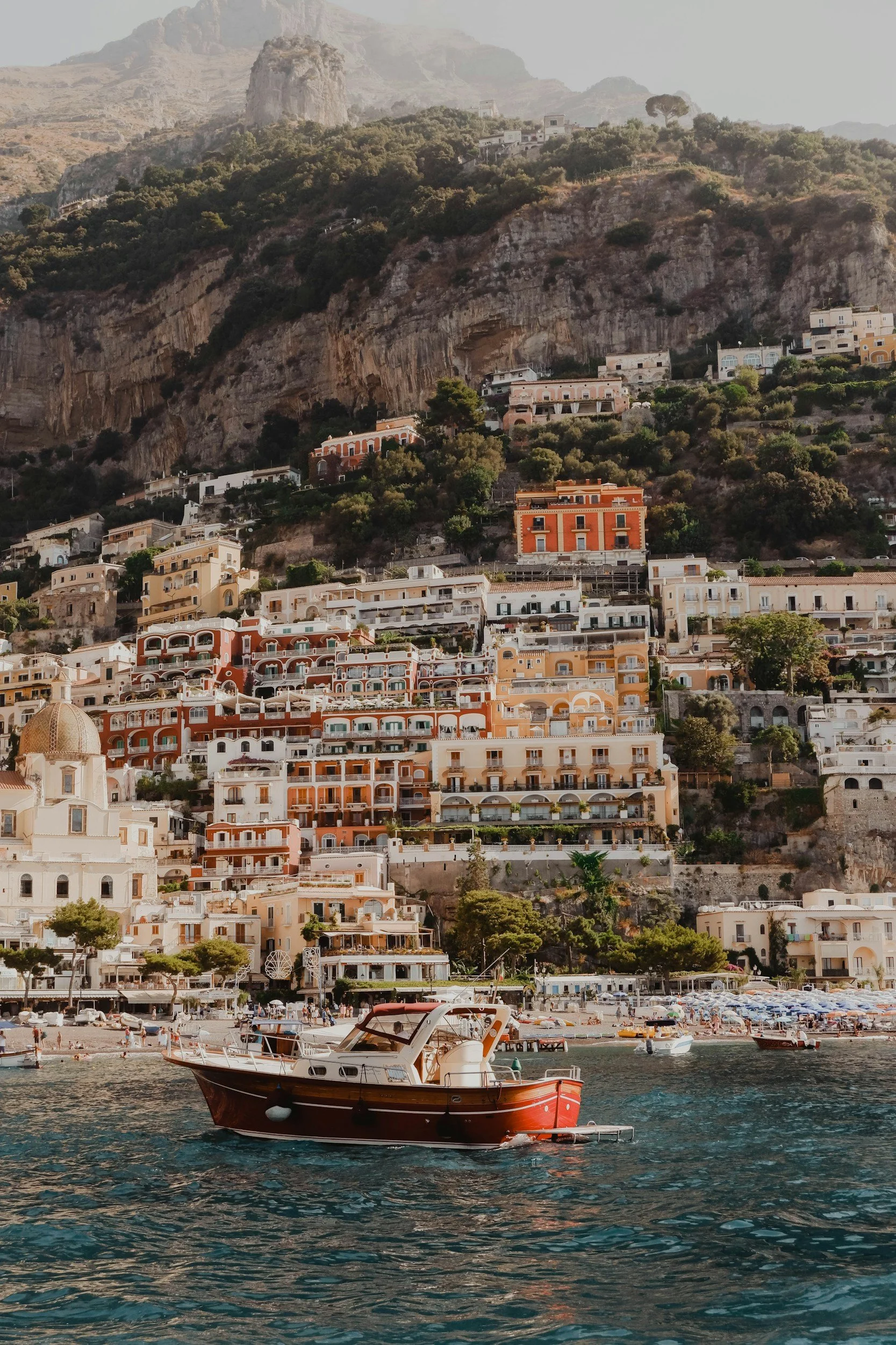 Coastal scene with colorful hillside buildings, a boat on the water, and mountains in the background.
