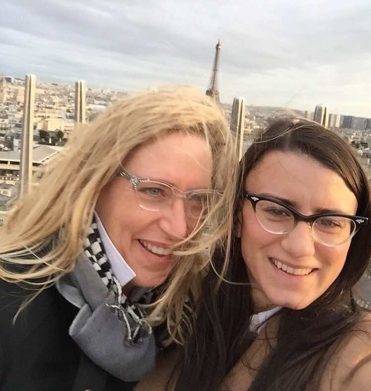 Two women smiling with the Eiffel Tower in the background on a cloudy day.