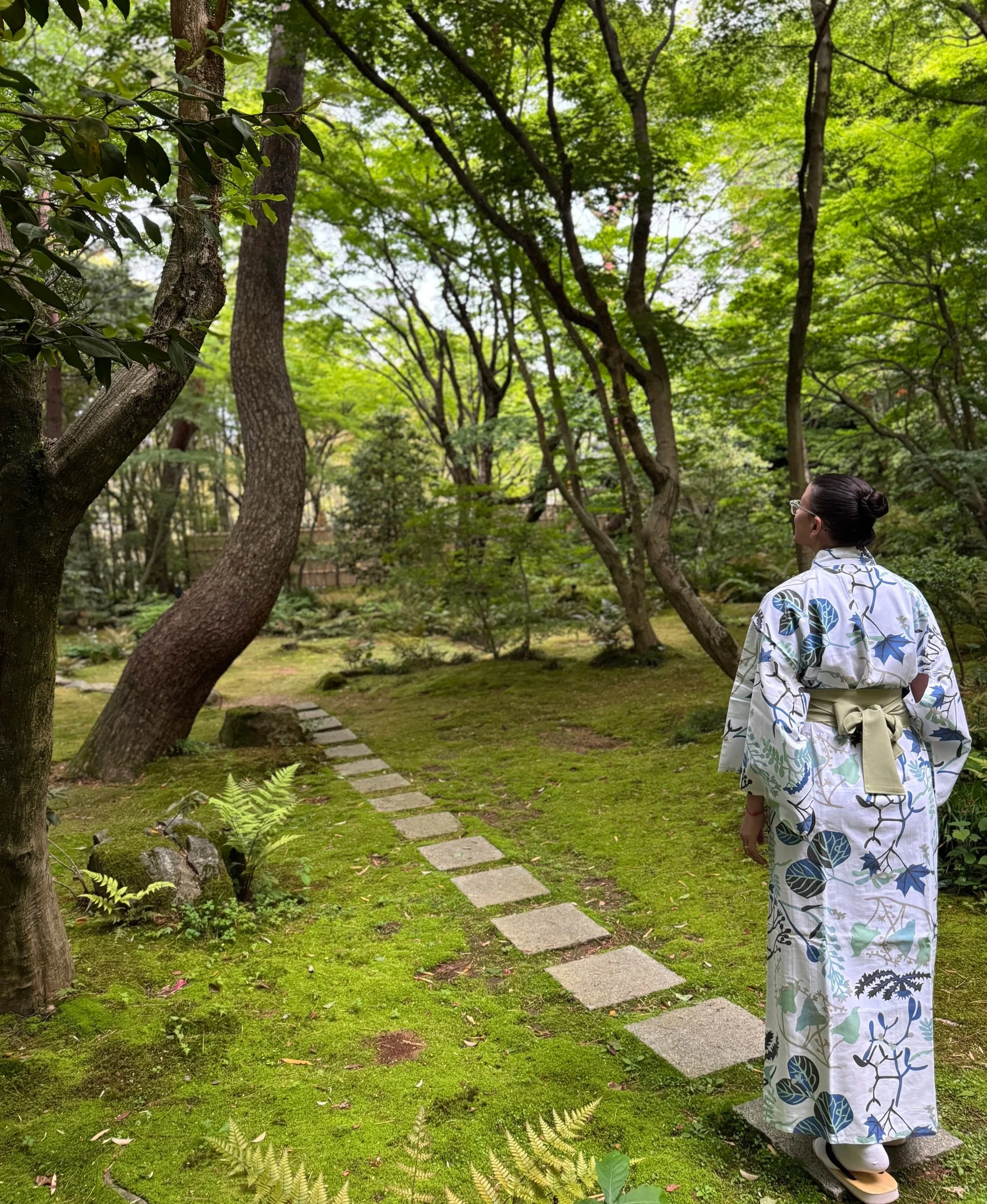 A person wearing a white yukata with blue and green leaf patterns walking through a lush Japanese-style garden with trees, ferns, and stepping stones.